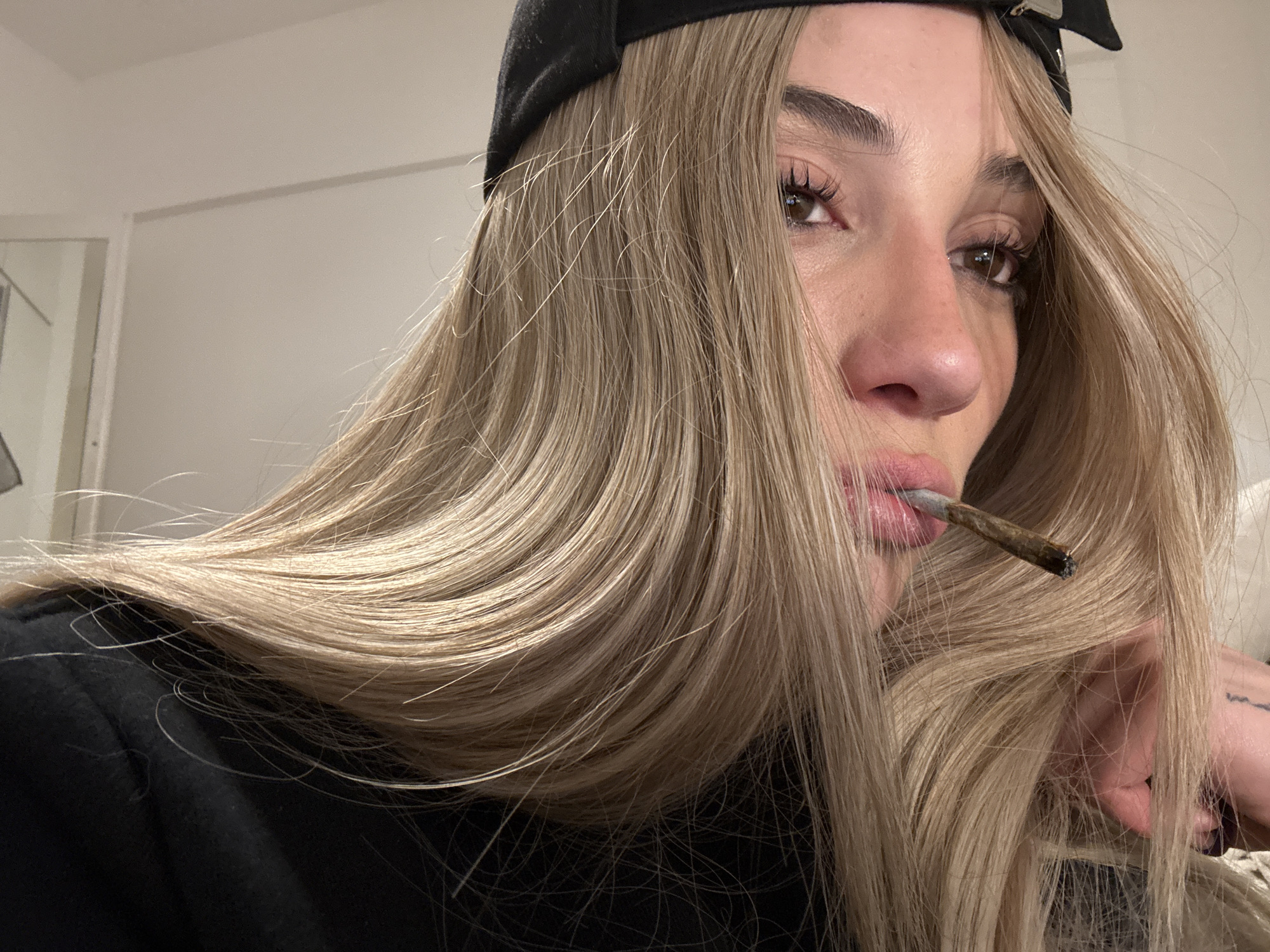 A fair-skinned woman with long, blonde hair, wearing a black baseball cap backwards, is holding a lit cigarette or joint in her mouth. She is wearing a black shirt and her right hand is visible near her face. The background is a plain, off-white wall. The location is not identifiable as Buenos Aires, Argentina based on the visual information provided.