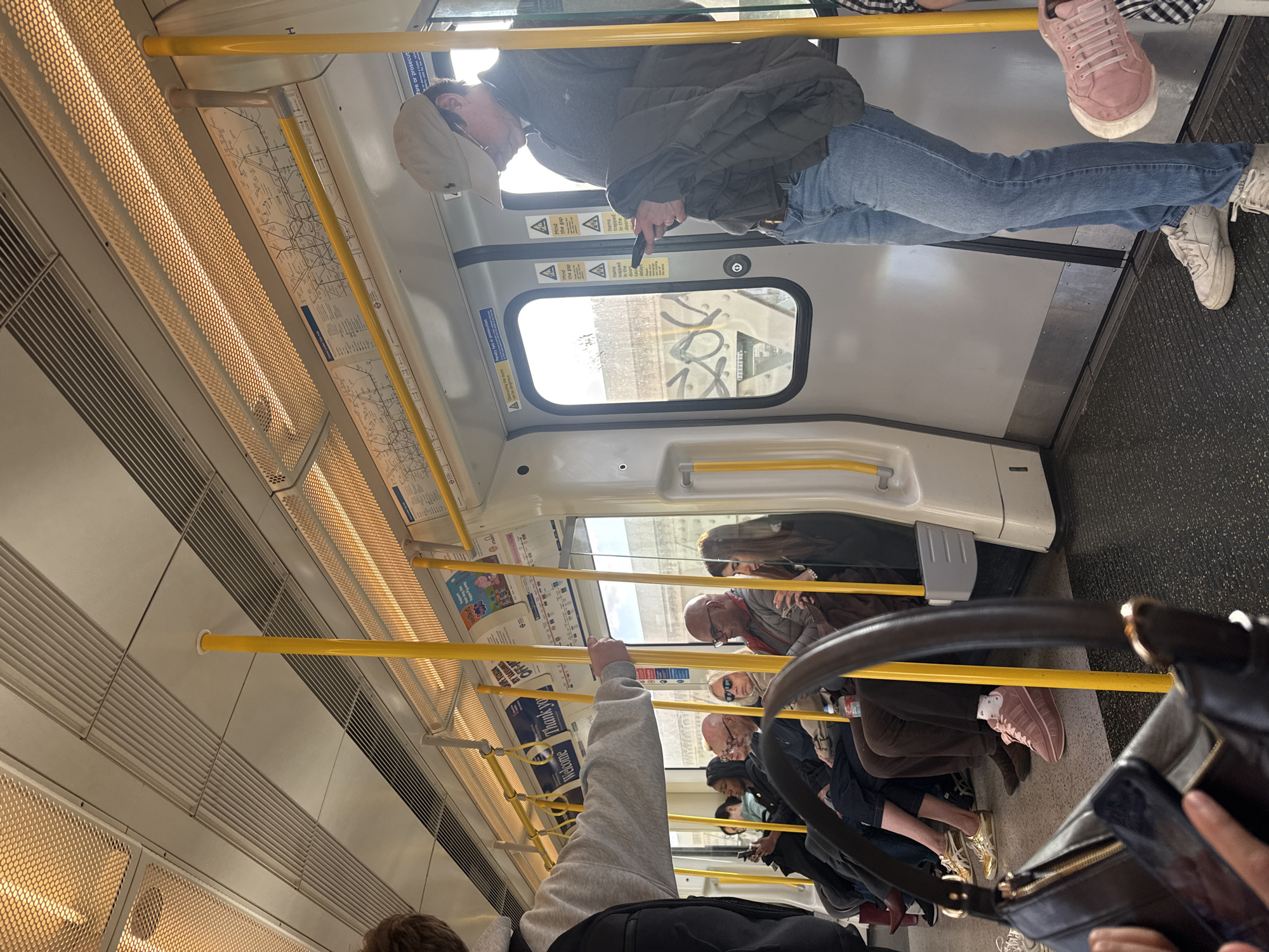 The image shows the interior of a train carriage, likely part of the London Underground, viewed from a slightly elevated, oblique angle. Several passengers are visible, either standing or seated, going about their journey during what appears to be daytime, judging by the bright light coming through the windows.
In the midground, a man wearing a beige cap, a grey zip-up jacket, and blue jeans stands near a closed train door, looking down at his phone. Below him, another person's legs are visible, clad in blue jeans and white sneakers, next to someone wearing pink sneakers. Further into the carriage, more passengers are seen, some holding onto the prominent yellow grab rails that span the length of the carriage. One person's arm is raised, gripping a yellow rail in the foreground.
The train's interior features distinctive overhead lighting panels with a perforated design, emitting a warm glow, alongside ventilation grilles. A London Underground map is visible on the wall above the windows, showing station names like "South Kensington" and "Victoria." The floor is dark and textured.
Visible text includes multiple instances of the warning "Mind the gap," as well as "Items trapped in the door" on the train door. Other partial texts like "Welcome Thank yo" and "Wellkid" (on an advertisement) are also present. Graffiti can be seen on an external structure through the window. The overall scene depicts a typical commute within a public transport system.