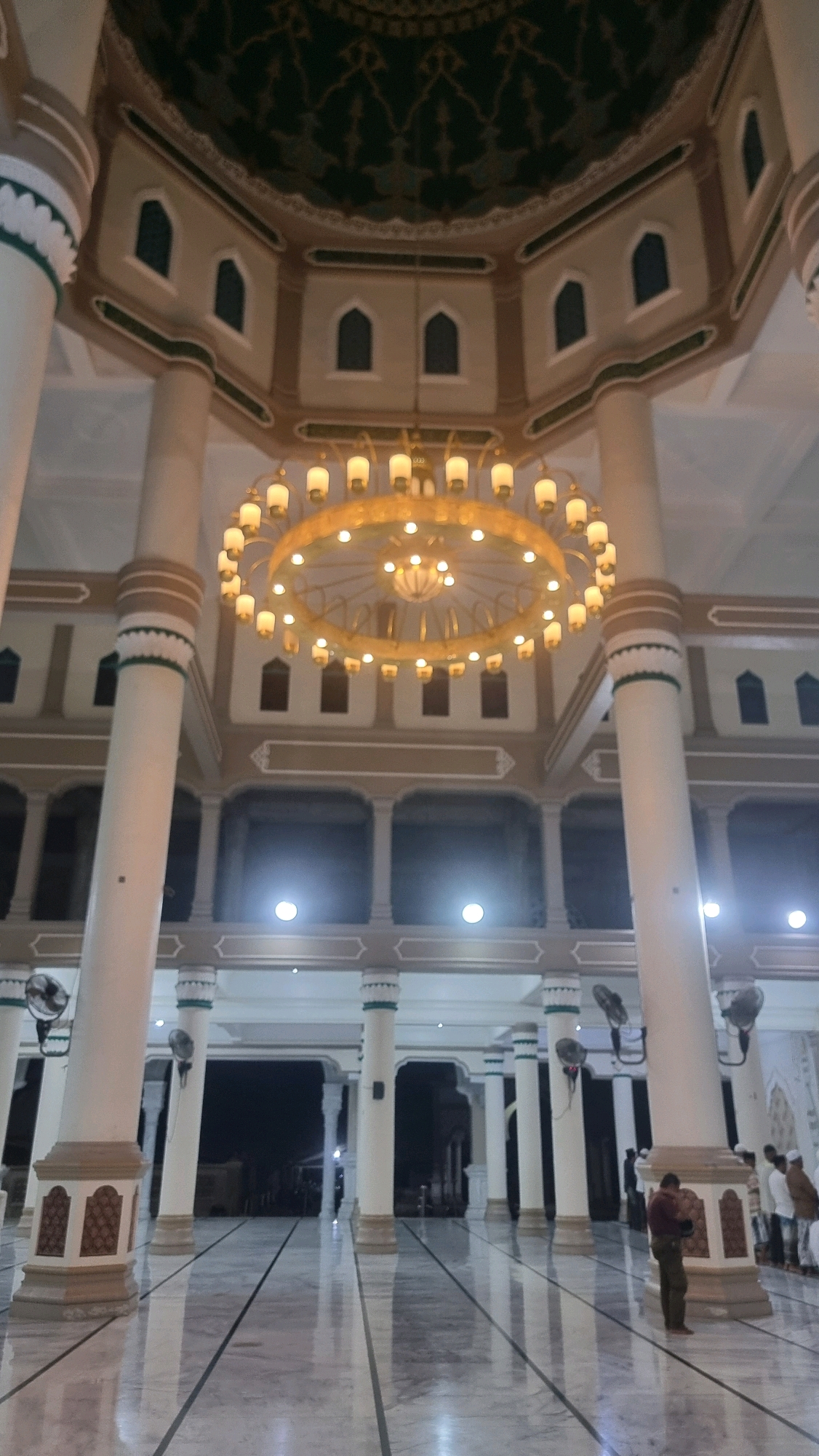 The image shows the interior of a mosque, likely the Islamic Center Lhokseumawe in Lhokseumawe, Indonesia, due to its architectural style and common features of mosques in the region. The central focus is a large, ornate chandelier with numerous lights hanging from the high, domed ceiling. The ceiling itself is decorated with intricate dark green patterns. Surrounding the chandelier are large, white, fluted columns with beige and green detailing that extend from floor to ceiling. Archways and windows are visible on the upper levels, with small arched windows set into recessed panels. The floor is polished marble with dark lines indicating prayer rows. In the foreground and mid-ground, several white columns and arched structures frame the scene. Fans are mounted on some of the columns. Towards the right side of the image, a few men are visible, some standing and others appearing to be praying. One man in the foreground is wearing a maroon shirt and khaki pants, standing with his hands clasped. The lighting is subdued, with the chandelier being the primary light source, creating reflections on the shiny floor. The overall impression is one of grandeur and solemnity.