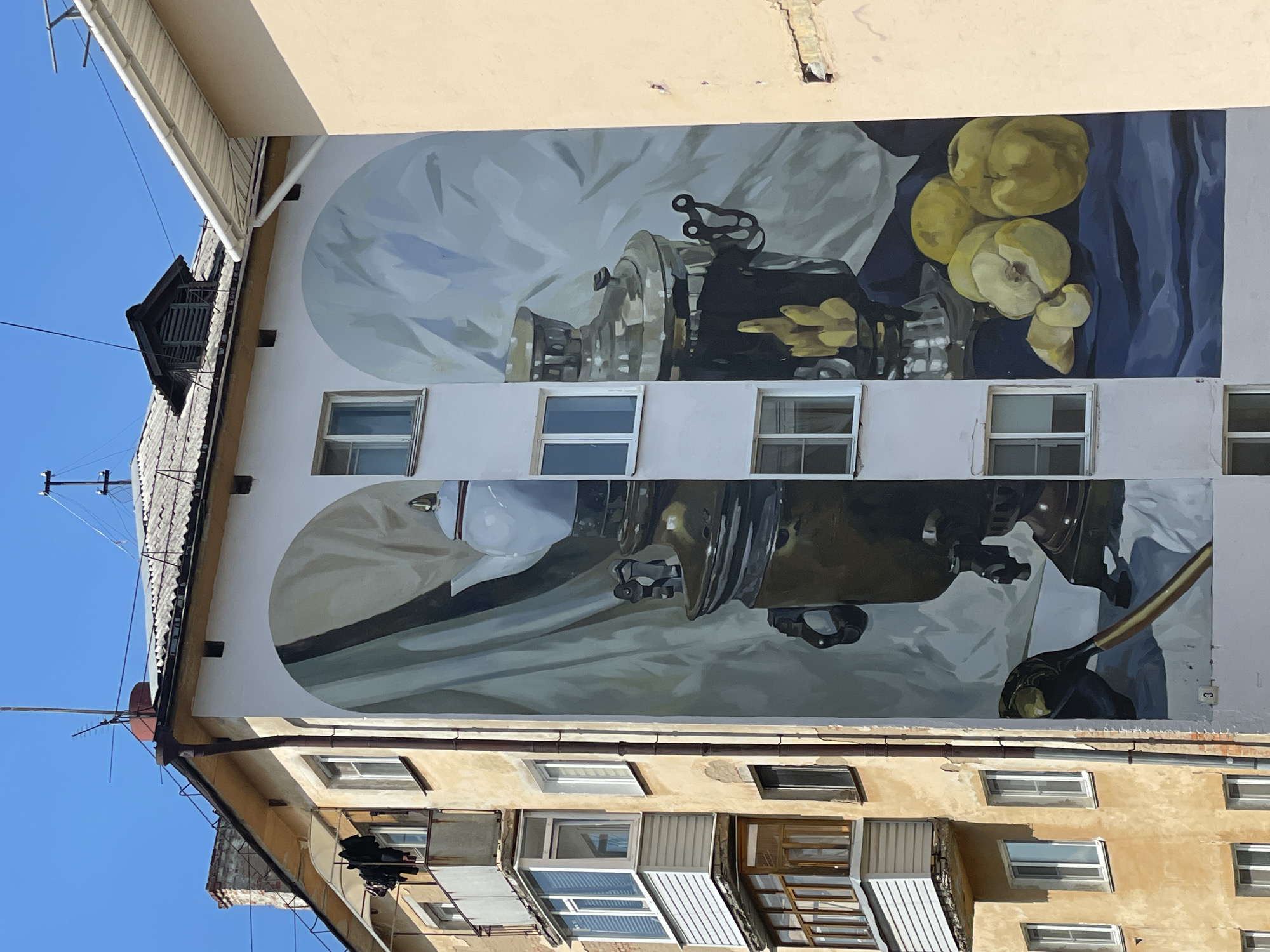 This is a photograph of a building located in Vologda, Russia, that features a large mural on its side. The mural depicts a still life composition with a prominent samovar, a teapot, and a pile of apples. The artwork is rendered in a realistic style, with careful attention to light and shadow. The building itself is a multi-story structure, likely an apartment building, with visible windows and balconies. The mural covers a significant portion of the building's facade, creating a striking visual element in the urban landscape. The surrounding environment appears to be a sunny day, with a clear blue sky visible above. The building's architecture suggests it is an older Soviet-era construction.