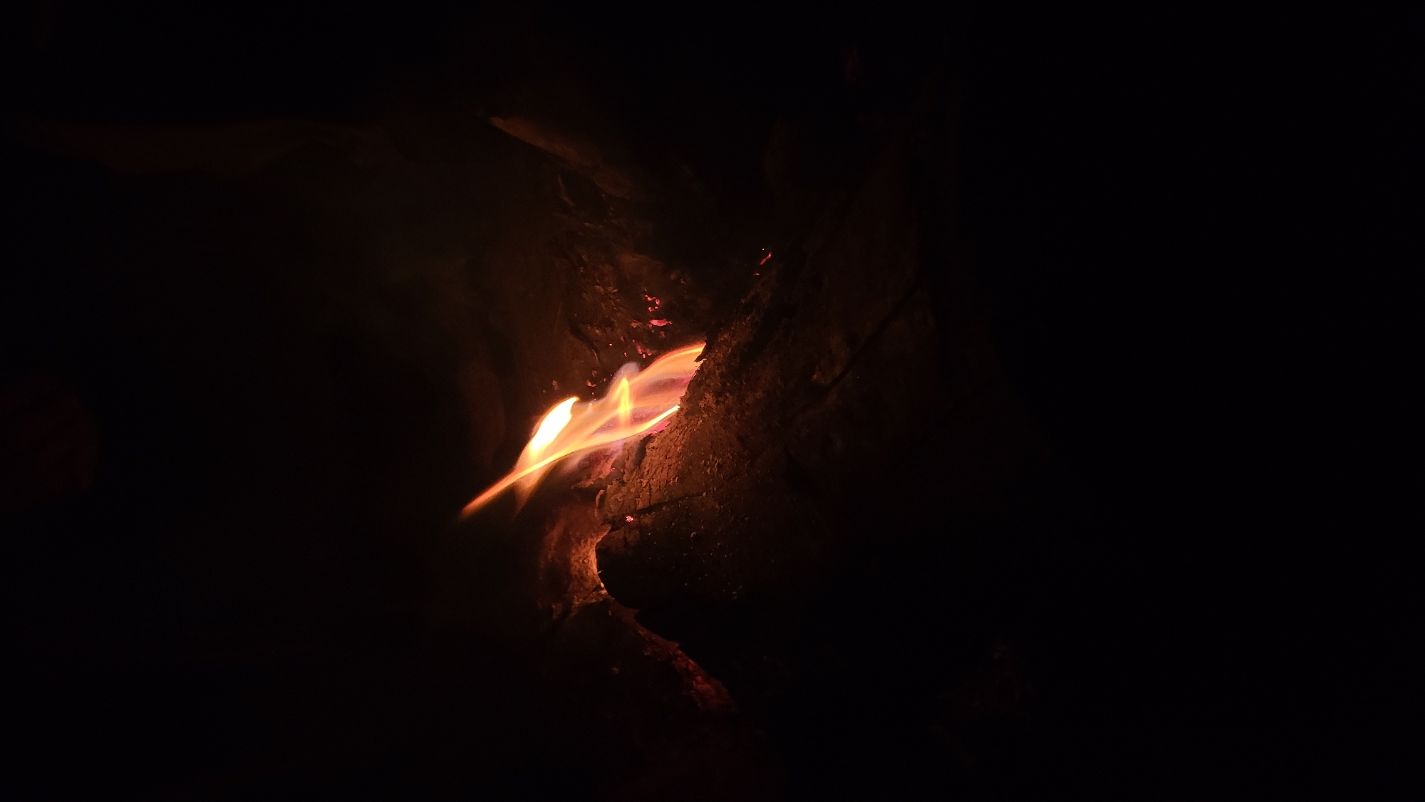 The media file presents a close-up, low-light photograph of a fire burning in a dark environment. The central subject is a vivid flame, glowing brightly with intense orange, yellow, and red colors. The flames appear somewhat blurred, suggesting either natural flickering, slight motion during capture, or a longer exposure time, creating streaks of light.
The fire is situated within or next to a dark, textured surface, most likely a log or piece of wood, which is partially illuminated by the firelight. This surface shows details of its rough texture, possibly bark or charred material, rendered in reddish-brown and dark tones. Scattered around the main flame are smaller, glowing red spots, indicative of embers burning intensely.
The background is predominantly dark black, with very little discernible detail, emphasizing the isolation and brightness of the fire. This suggests the photograph was taken at night or in a very dark setting, where the fire is the primary light source. The overall impression is one of warmth and elemental beauty against the surrounding darkness. Given the location context of Khir Ganga, India, this could be a campfire or a fire for warmth in a natural, possibly mountainous or trekking environment. There are no people, other distinct objects, or visible text present in the image.