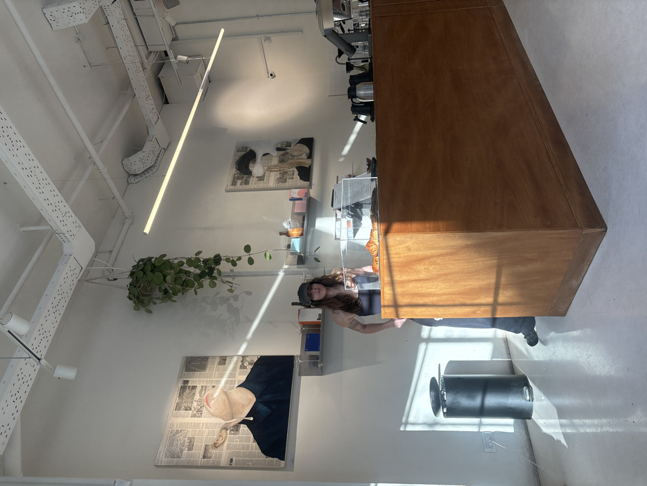 An interior view of a contemporary cafe or bar in Tel-Aviv, Israel, features a single female individual standing behind a long wooden service counter. The woman, identified as staff, wears a dark baseball cap, a dark tank top, and dark trousers; tattoos are visible on her right arm. She stands upright, facing the viewer, with her hands positioned near her waist.
The environment is characterized by white walls and a white ceiling with exposed industrial-style cable trays and track lighting. Bright, directional sunlight enters from the left, casting distinct diagonal shadows across the white floor and lower wall, partially illuminating the woman and the counter.
On the wooden counter, a clear acrylic display case holds several croissants. Behind it, various items of coffee preparation equipment, including multiple dark pitchers and a portion of an espresso machine, are visible. The walls display two large framed artworks; the left depicts a person's chin and neck looking upward with an object in their mouth against a newspaper collage, while the right features a face partially obscured by a white cloud-like shape, also against a newspaper background.
Below the artworks, minimalist metal shelves hold bottles and small boxes. A potted plant with long green vines hangs from the ceiling between the two artworks. A long, linear LED light fixture is suspended horizontally from the ceiling. In the lower-left, a black cylindrical pedal bin with its lid slightly ajar stands on the floor. A small white security camera is mounted high on the right wall.