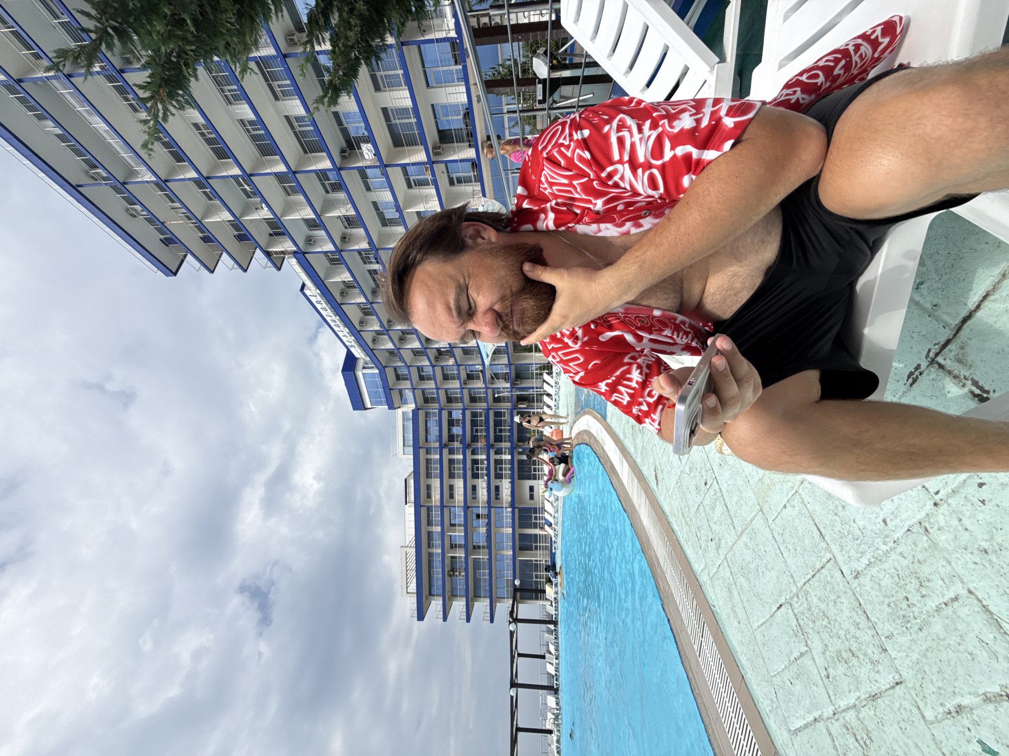 A man with a beard, wearing a red and white patterned shirt and black shorts, sits on a white lounge chair next to a swimming pool. He is holding a smartphone in his left hand and resting his right hand on his chin. In the background, a large, multi-story hotel with blue accents is visible, along with a swimming pool filled with blue water and several people. The sky is cloudy. The scene appears to be at a resort or hotel with a pool in Sevastopol, Ukraine.