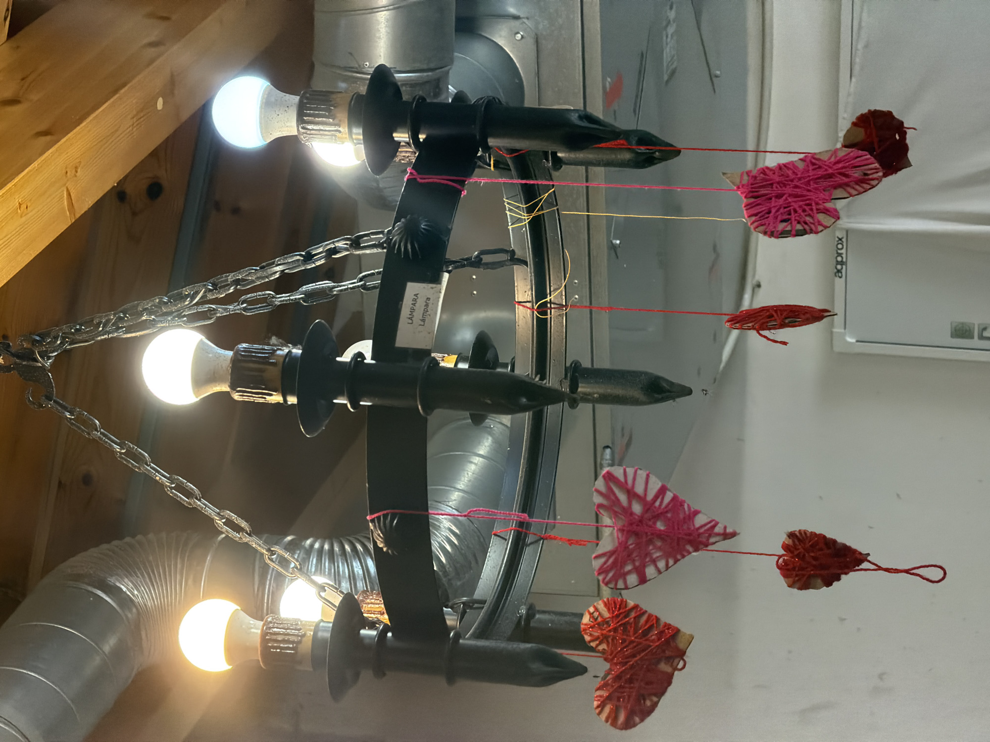 A black wrought iron chandelier is suspended from a wooden beam and metal ducts in an indoor space, possibly a workshop or unfinished room, in Valdefresno, Spain. The chandelier features six candle-style light fixtures, each fitted with an LED bulb emitting a bright white light. The chandelier is adorned with several handcrafted heart-shaped ornaments made of cardboard and wrapped with red and pink yarn. These ornaments are suspended from the chandelier by thin red and yellow threads. One heart is larger and more intricate, while others are smaller and simpler. In the background, the metallic gleam of air ducts and wooden ceiling beams are visible, suggesting a utilitarian or construction environment. A white label with the text "LÁMPARA" is affixed to the chandelier. A white refrigerator or cabinet with the brand name "aqprox" is partially visible in the lower right of the image. The overall scene is dimly lit, with the primary illumination coming from the chandelier's bulbs. There are no people present in the image.