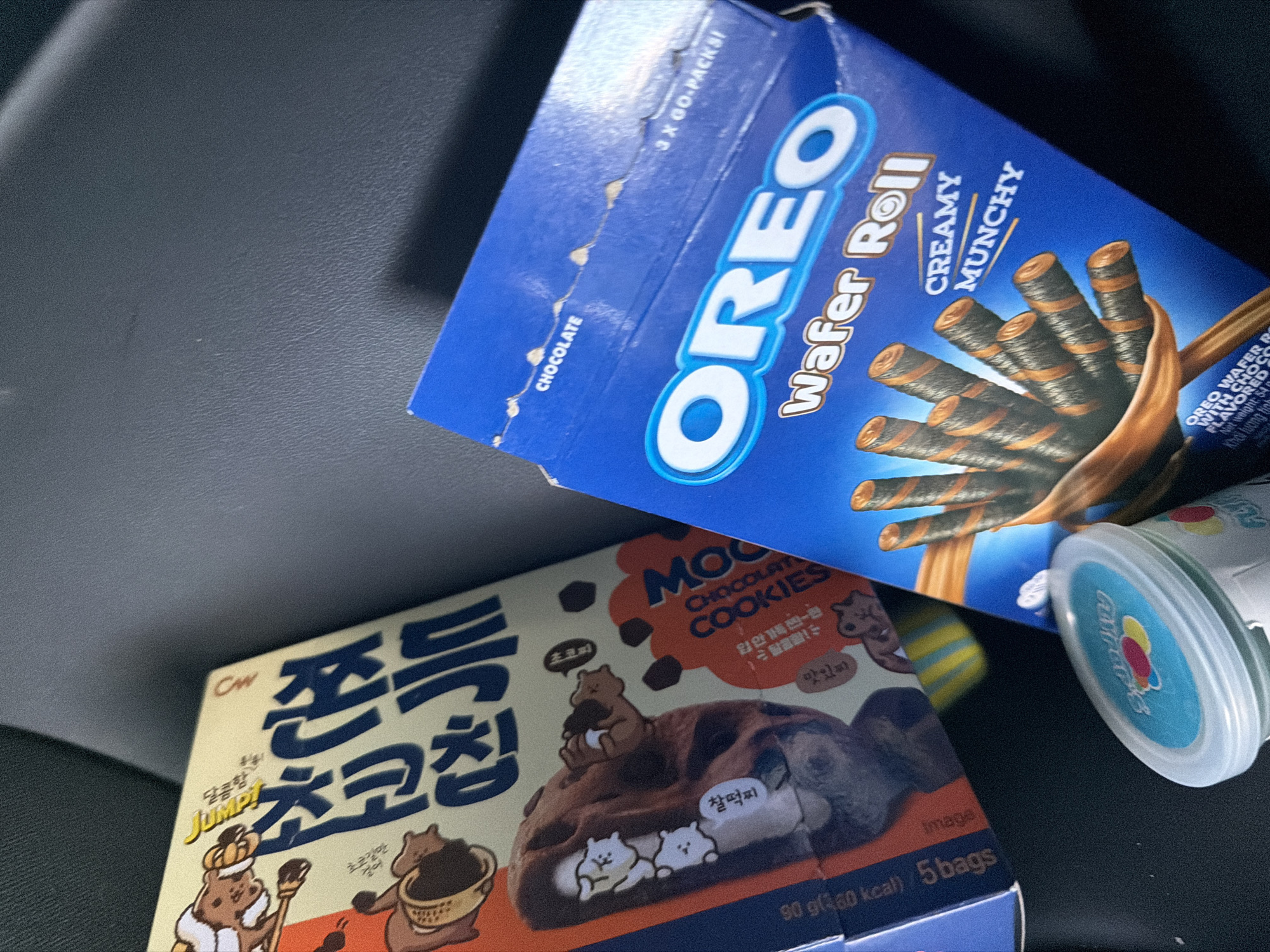The image shows a close-up of several snack food items on a dark, textured surface, likely the interior of a vehicle. In the foreground, a partially visible blue box of "OREO Wafer Roll" with "CREAMY MUNCHY" written on it is prominent. Inside the box, several chocolate-striped wafer rolls are visible, with a CGI depiction of a hand holding them. To the left of the Oreo box is a light-colored cardboard box with Korean text and cartoon bear imagery, featuring what appears to be "MOO CHOCOLATE COOKIES" written in orange text. A small, clear plastic container with a white lid and a colorful label is also visible in the lower part of the image. The lighting suggests an indoor setting, possibly the interior of a car. There is no indication of the location being in Ödsmål, Sweden; this is a generic still life of snacks.