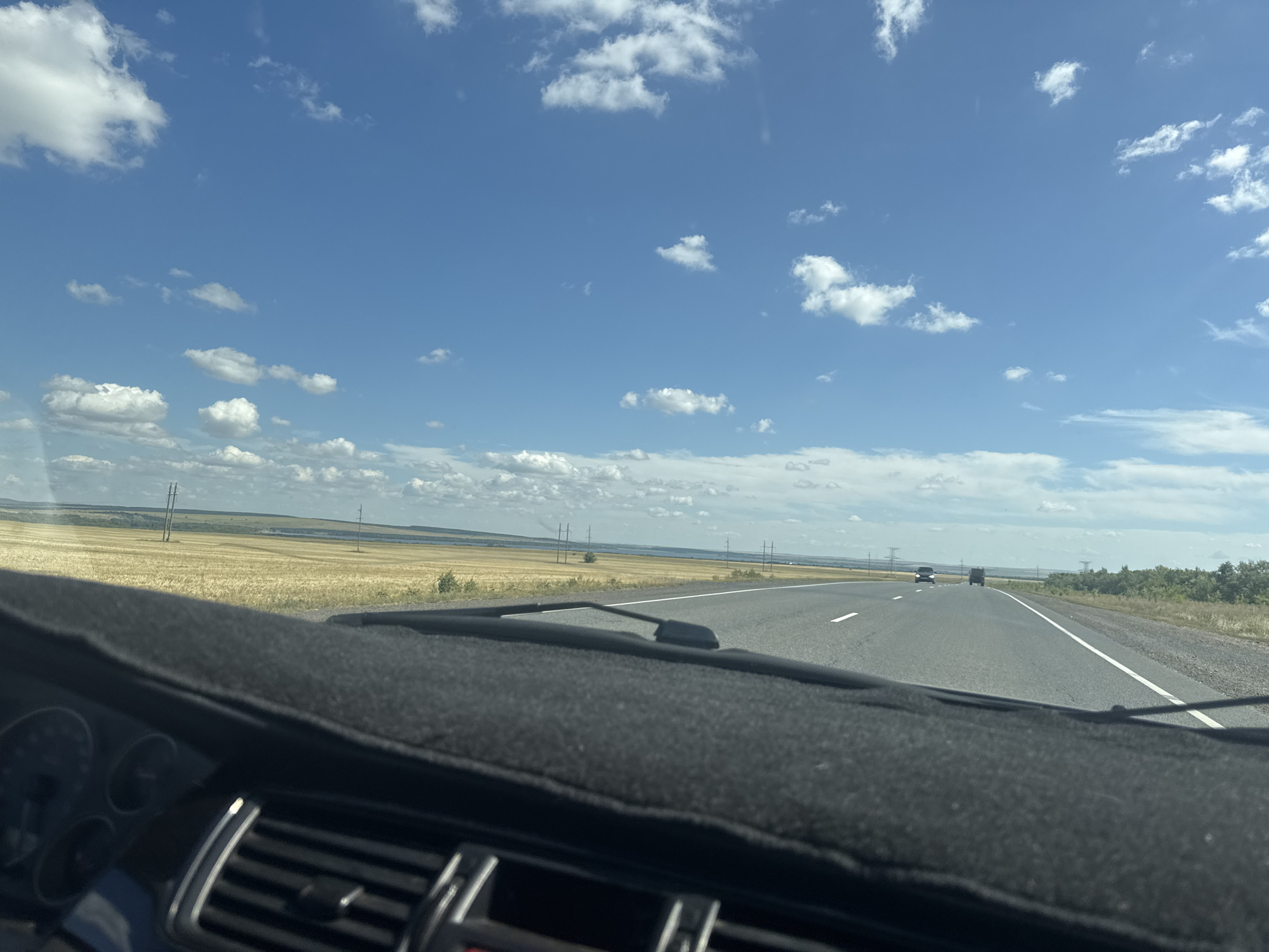 The image shows a view from the dashboard of a car driving on a highway. The road is asphalt and has white lane markings. In the distance, there are fields of dry grass, power lines, and trees. The sky is blue with scattered white clouds. There are a few other vehicles on the road, including a truck and a car. The foreground of the image is dominated by the car's dashboard, which is covered with a black fabric mat. The dashboard has some visible vents and parts of the instrument panel. The scene appears to be a sunny day in a rural or semi-rural area.