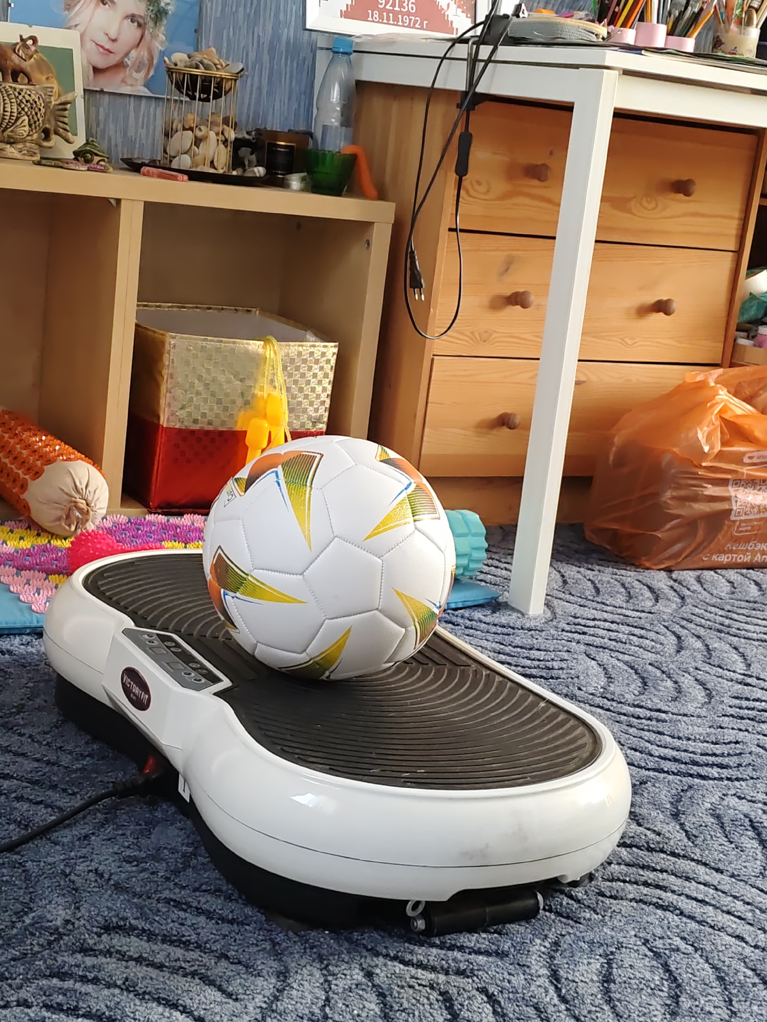 The image depicts a domestic indoor scene, likely a living room or bedroom, with a focus on fitness equipment and decorative items. In the foreground, a white vibration platform, identified by the "VICTORYFIT" logo, is positioned on a blue, textured rug. A soccer ball with a multicolored geometric pattern rests on the platform. To the left of the platform, a cylindrical orange object with a textured surface, possibly a massage roller, is visible.
Behind the platform, a light-colored wooden shelf unit holds various objects. On the top shelf, a framed picture of a woman with blonde hair is displayed, along with a decorative cage filled with stones and other small items. A clear plastic water bottle is also present on this shelf. Below, a red and gold patterned box is placed on the lower shelf.
To the right of the shelf unit, a light-colored wooden dresser with three drawers is visible. A white table or desk is positioned in front of the dresser, with a black power cord extending from it. An orange plastic bag is placed on the floor to the right of the table.
The overall environment suggests a residential space where fitness and personal interests are integrated. There are no people visible in the image. The specific location within the city of Rostov-on-Don, Russia, cannot be determined from the visual information provided.