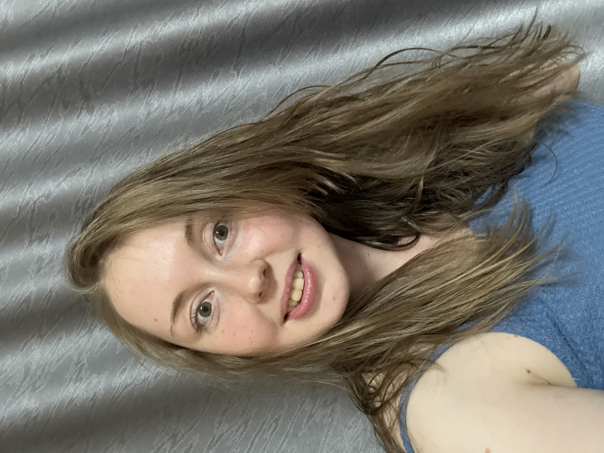 A woman is pictured from the chest up, looking at the camera. She has light brown hair that falls around her face and shoulders. She is wearing a blue top. Behind her is a grey, textured curtain.