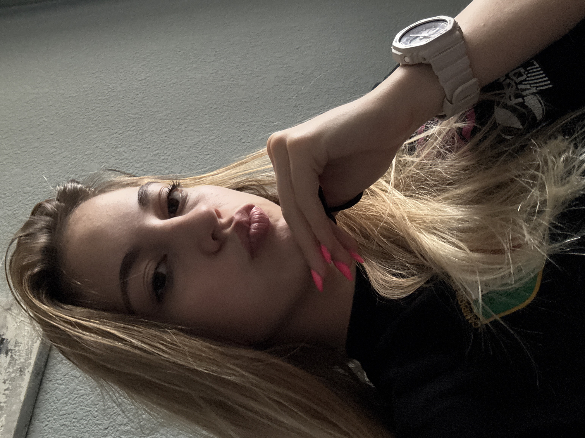 A young woman with blonde hair is lying down, facing the camera at an angle. She has fair skin, dark eyes, and is pursing her lips. She is wearing a black top, and her right arm is raised with her hand near her face. She has pink nail polish. A light gray watch is visible on her wrist. The background appears to be a light gray wall. The image does not provide enough information to determine a specific location in Sofia, Bulgaria, or to confirm any actions or interactions.