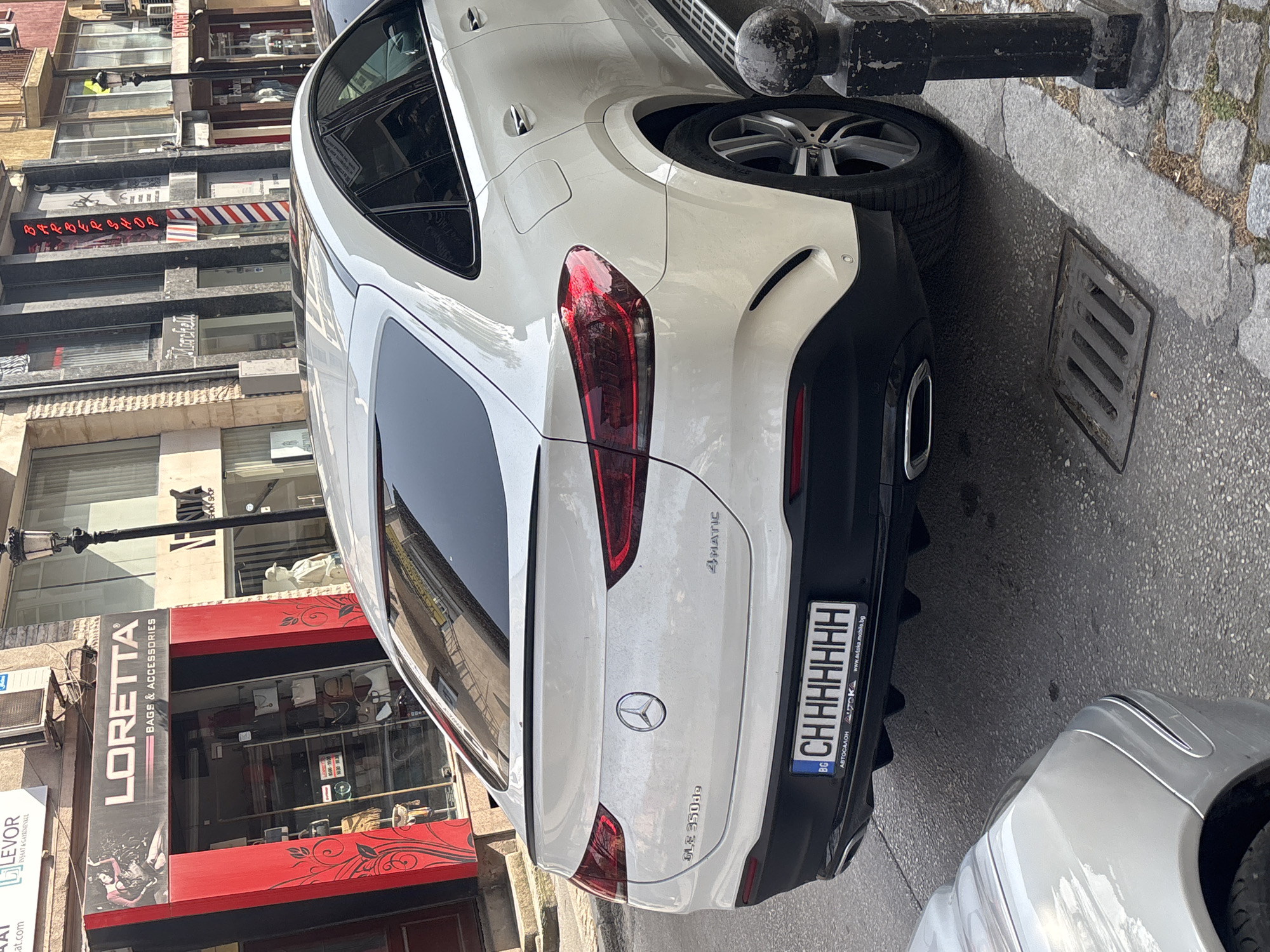 The image shows the rear of a white Mercedes-Benz GLE 350de 4MATIC car parked on a city street. The car has a Bulgarian license plate reading "CHHHHHHH". The background features buildings, including a shop called "LORETTA BAGS & ACCESSORIES" with a red storefront and a sign for "BARBERSHOP". The street appears to be in an urban area, with elements like a bollard and a drainage grate visible. The location is Plovdiv, Bulgaria, based on the URL on the license plate, "www.autoka.mobila.bg". Another car is partially visible in the bottom right corner of the image.