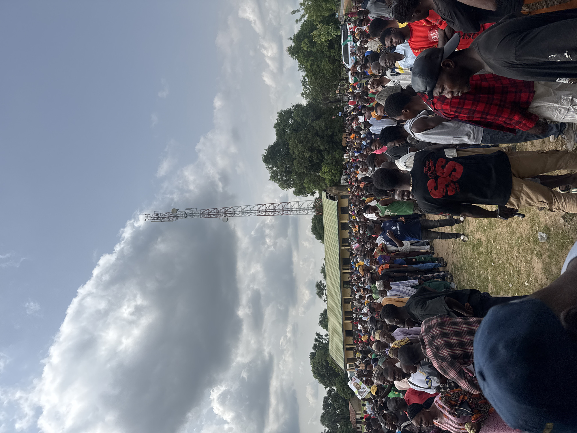 The image shows a large crowd of people gathered outdoors in Akwanga, Nigeria. The people are densely packed together, facing in the same direction. Some individuals are standing while others are seated or kneeling on the ground. The crowd is predominantly made up of African men. A communications tower is visible to the left of the crowd, against a sky filled with dramatic clouds. A green roof can be seen in the background, behind some trees.