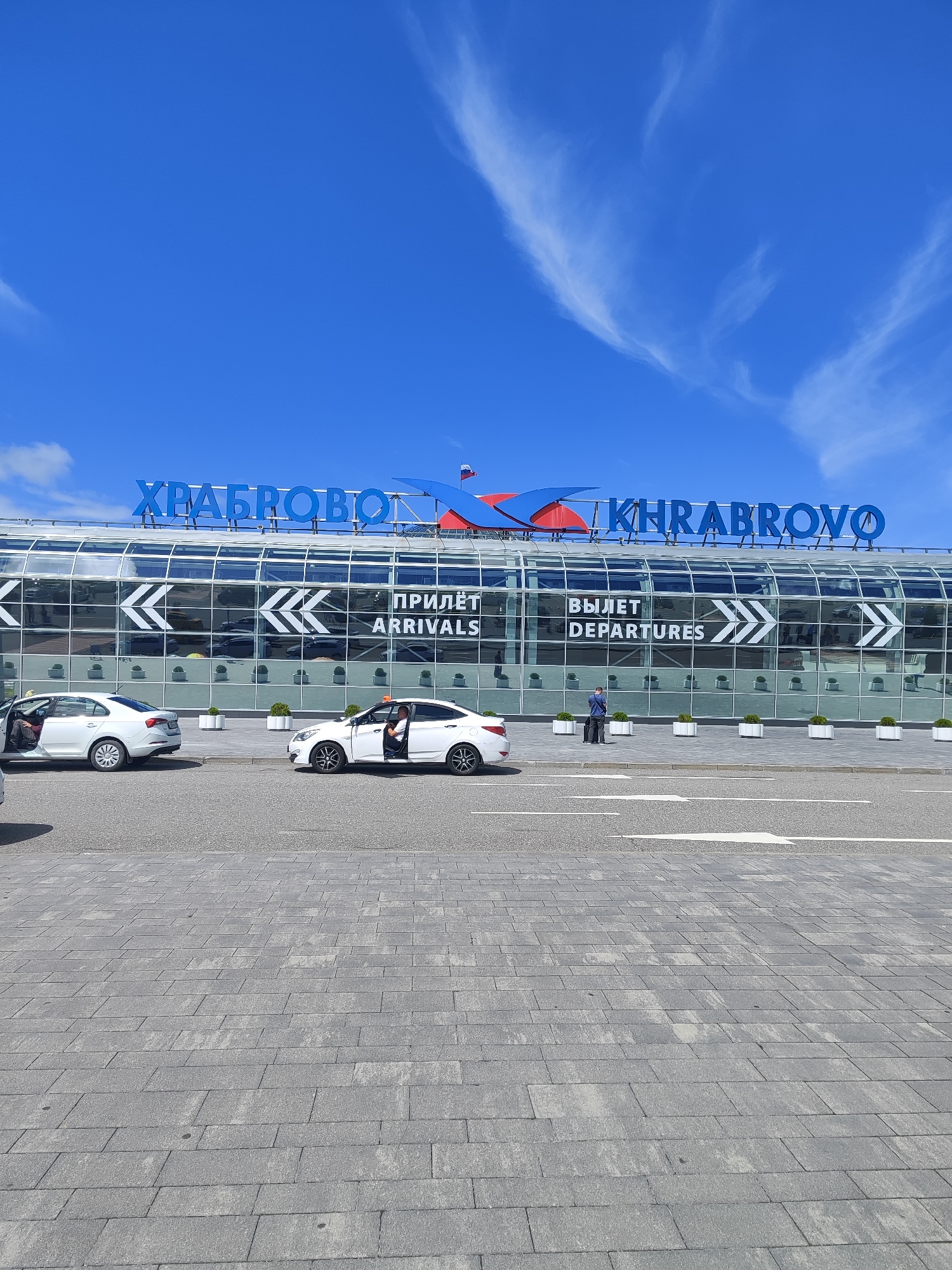 The image shows an exterior view of Khrabrovo Airport, located in Kaliningrad Oblast, Russia. The airport's terminal building features a modern glass facade with large blue lettering spelling out "KHRABROVO". Above the entrance, two directional arrows are visible, with text indicating "ПРИЛЕТ ARRIVALS" on the left and "ВЫЛЕТ DEPARTURES" on the right. Two white sedans are parked on the asphalt in front of the building, with their doors open. One car has a driver inside, while the other appears to be occupied by passengers. A person in a blue shirt and dark pants stands between the cars, holding luggage. The ground in the foreground is paved with grey interlocking bricks, and white directional arrows are painted on the asphalt road. The sky above is a vibrant blue with wispy white clouds.