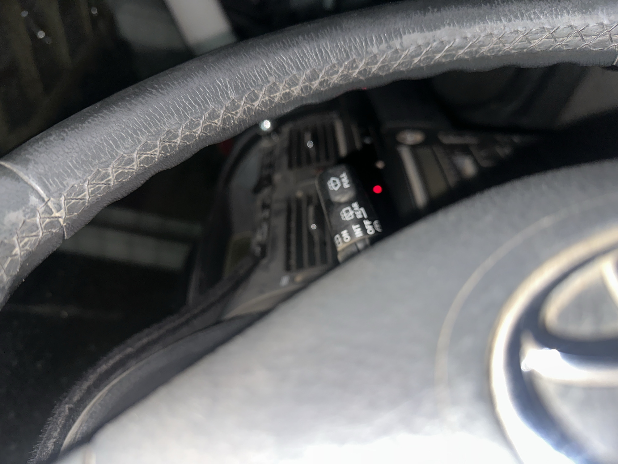 The image provides a close-up view of a car's interior, focusing on the steering wheel and a portion of the dashboard. The steering wheel, upholstered in dark leather with visible stitching, occupies a significant part of the frame. To the left of the steering wheel, a set of controls for the windshield wipers is visible, featuring settings such as "OFF," "INT," "ON," and "REAR," along with a button labeled "FULL." A small red light is also illuminated near these controls. Behind the steering wheel and controls, the dashboard is partially visible, including an air vent. The overall impression is of a car parked indoors, possibly in a garage or covered parking area, given the ambient lighting and background elements. The image does not depict any people or specific street views that would allow for precise localization within Тайгараев, Kyrgyzstan.