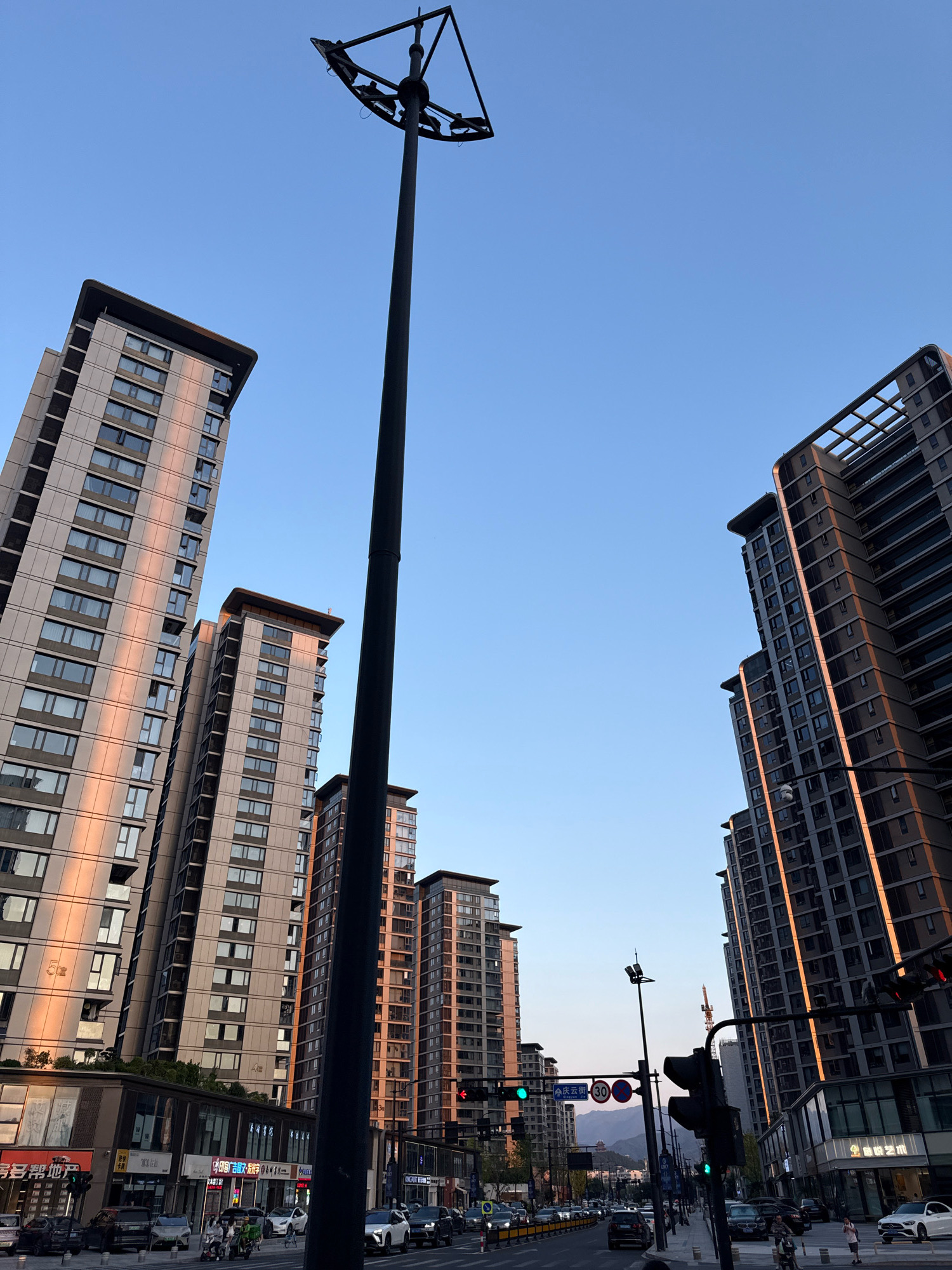 This is a photograph taken at eye level, looking up at several modern apartment buildings and a street scene at dusk in Yiwu, China. The sky is a clear, pale blue. A tall, dark street lamp with multiple lights is positioned in the foreground, slightly off-center to the left. Two large apartment buildings dominate the left side of the frame, their facades illuminated by the warm, golden light of the setting sun, highlighting the vertical lines of windows. To the right, another cluster of similar apartment buildings, also in shadow and catching the last rays of light, rise towards the sky.
Below the buildings, a street is visible with moderate traffic. Several cars, a few scooters, and pedestrians are present. Traffic lights are visible, showing a green light. Street signs with Chinese characters are also present. In the distance, behind the buildings, the outlines of mountains can be seen against the horizon. The overall impression is of a bustling urban environment at the end of the day.
