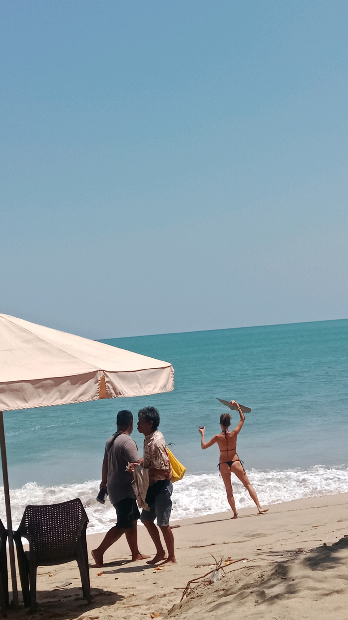 The image captures a sunny day at Kuta Beach, Indonesia. In the foreground, a beige beach umbrella casts a shadow over a black plastic chair. To the right of the umbrella, two men are standing on the sand, appearing to be in conversation. One man, on the left, wears a grey t-shirt and dark shorts, and is holding a dark object in his right hand. The other man, to his right, is wearing a patterned short-sleeved shirt, dark shorts, and has a yellow bag slung over his shoulder. He is holding a white cloth in his left hand.
Further in the background, towards the turquoise ocean, a woman wearing a black bikini is standing with her back to the camera. She is holding a flat, light-colored object above her head with her right hand and a glass in her left. White waves are breaking on the sandy shore. The sky is clear and blue. The overall scene depicts a leisurely day at the beach.