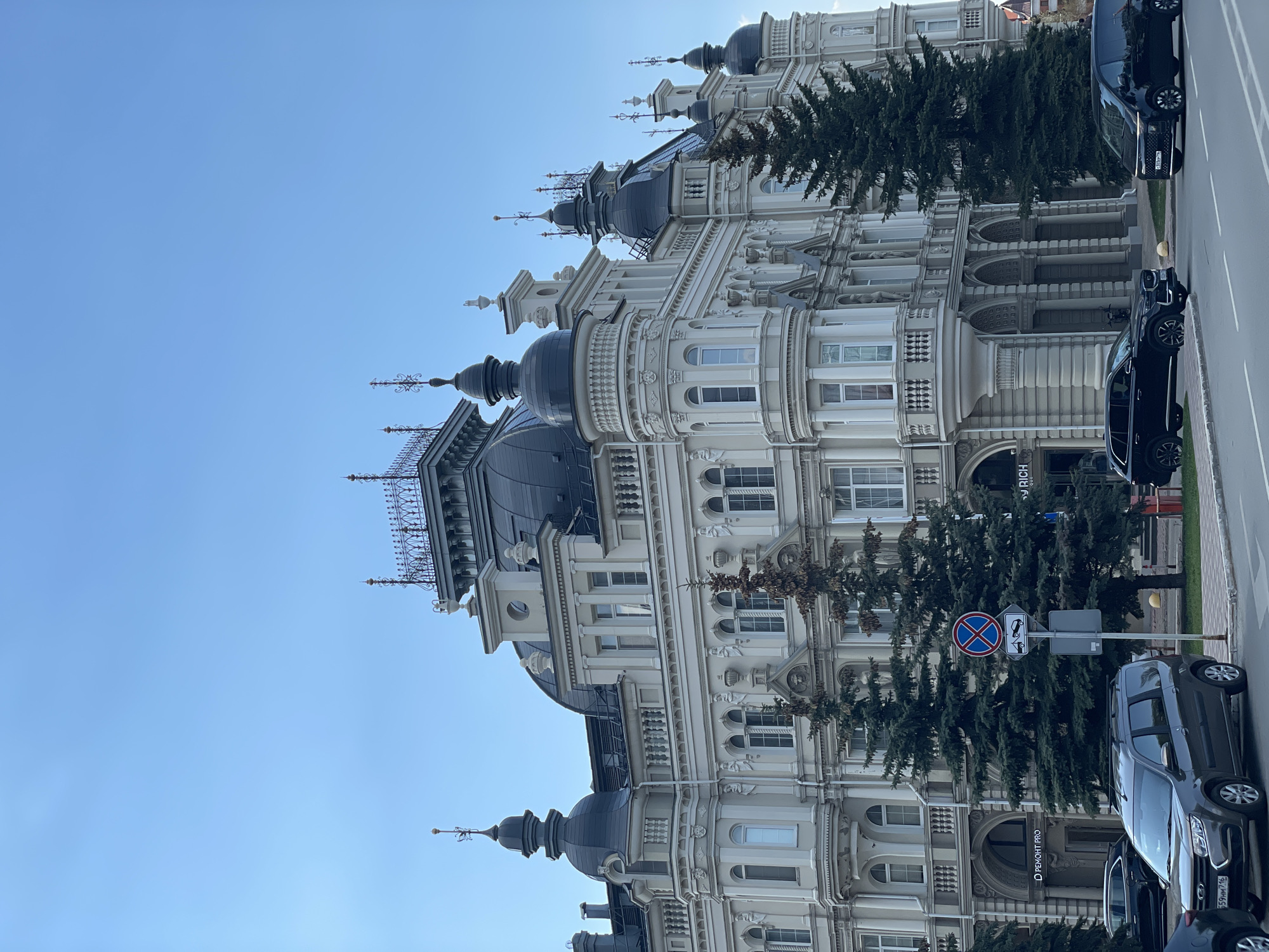 The image shows a building with a European-style architectural design, featuring multiple domes, intricate facades, and decorative elements. The building is primarily white or light-colored, suggesting a classic aesthetic. The image is oriented vertically, with a clear blue sky in the background. A few evergreen trees stand near the building, partly obscuring the lower portion. In the foreground, there are several parked cars along a road, indicating an urban setting. A "no parking" sign is also visible. The location appears to be in Kazan, Russia.