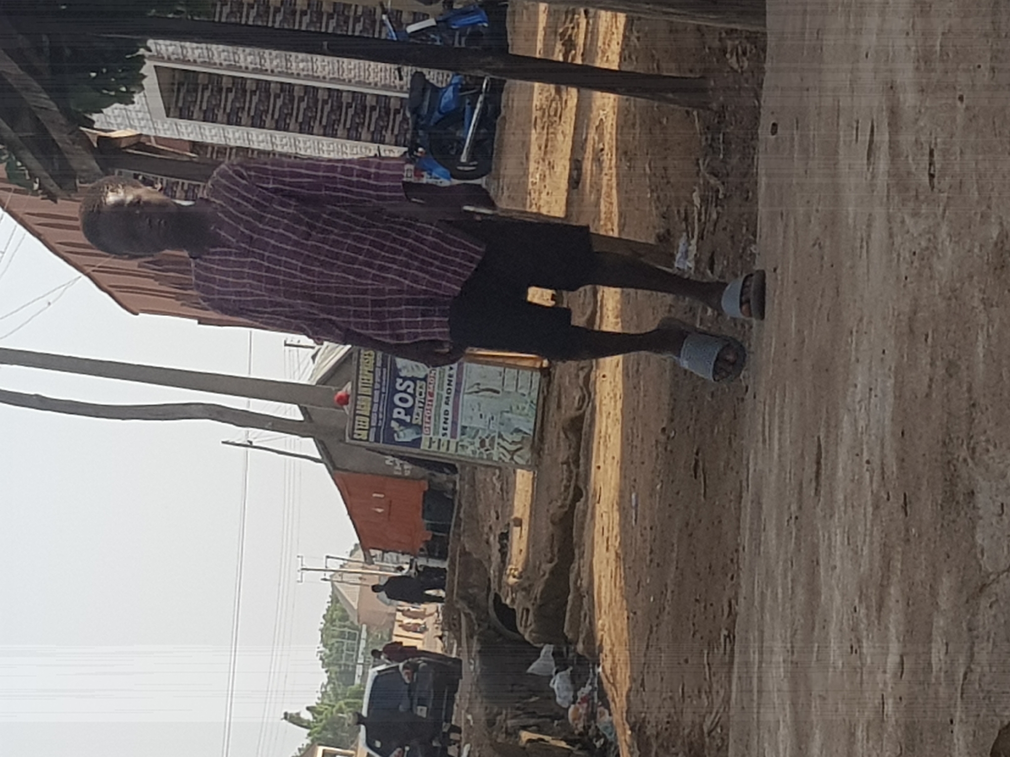 A full-body shot of a young Black male is captured in an outdoor setting, likely a street in Kufena, Nigeria. The individual is wearing a red and white plaid long-sleeved shirt and black shorts, with gray slide sandals on his feet. He is walking towards the viewer, with his head slightly bowed and his gaze directed downwards. His posture suggests a casual, perhaps pensive, demeanor.
The environment appears to be a dirt road with uneven terrain, characterized by loose soil, rocks, and some scattered debris, including plastic waste on the left side of the frame. In the background, several structures are visible. To the left, a dark-colored car is parked, and a few other individuals are present, though less distinct. A red cargo truck is also visible, with a sign on its side displaying "POS SERVICES," "DEPOSIT MONEY," and "SEND MONEY." Behind these elements, a building with a textured facade and what looks like a metallic roof is present. The sky is bright, suggesting daytime, with a slightly hazy or overcast condition. Utility poles and overhead wires are also part of the background landscape, indicating an inhabited area. The overall impression is that of a typical street scene in a town or city.