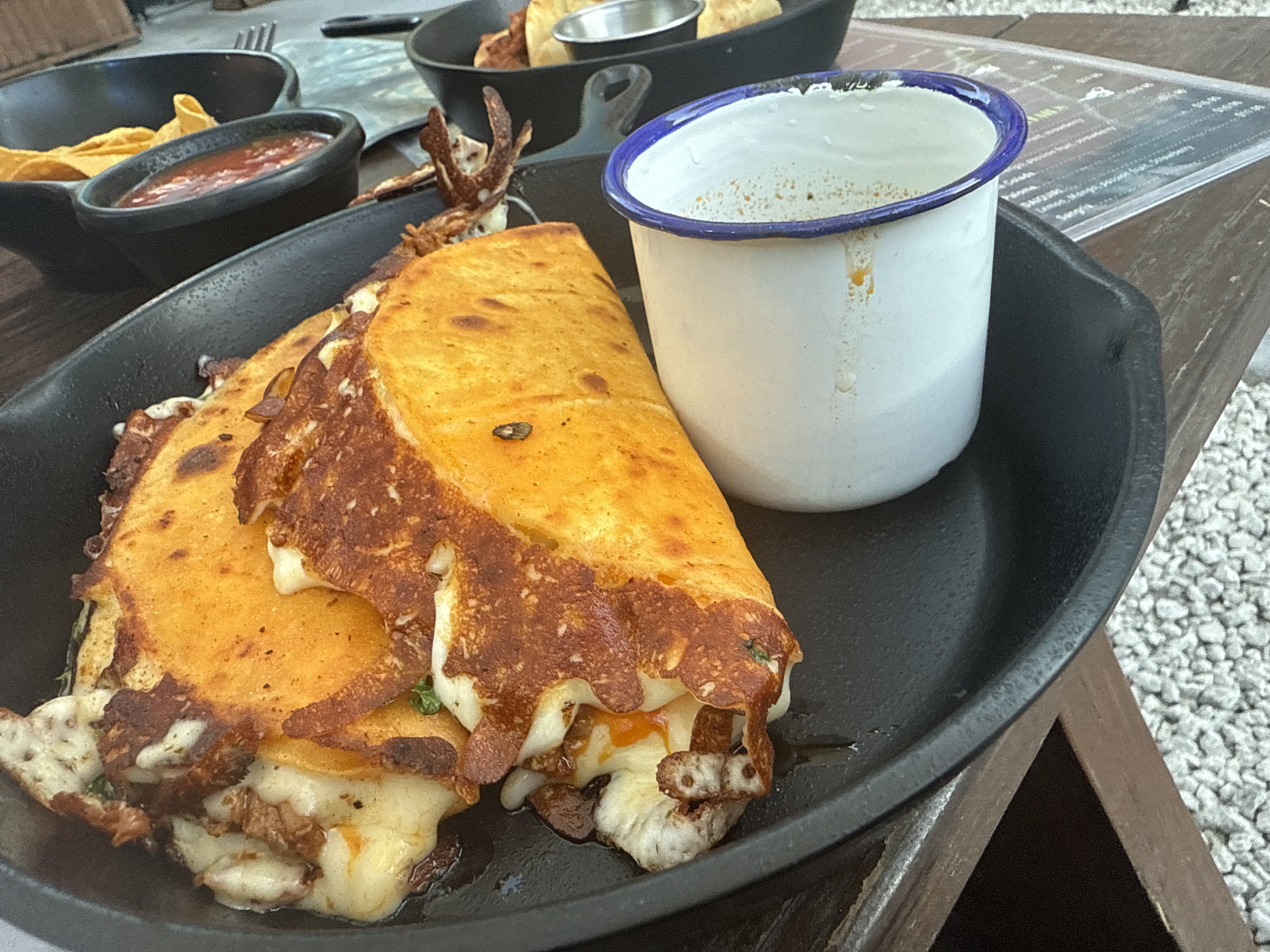 A close-up shot reveals a black cast-iron skillet filled with two golden-brown quesadillas oozing with melted cheese. The quesadillas are next to a white mug with a blue rim, possibly containing a beverage. In the background, other food items are visible in black bowls or pans, including chips, salsa, and other dishes. A menu card is also subtly visible, suggesting a restaurant setting. The shot appears to be taken outdoors, on a wooden table. Based on the style of the food and restaurant presentation, this could be a trendy or upscale dining establishment, potentially located in a city like Hollywood, United States of America.