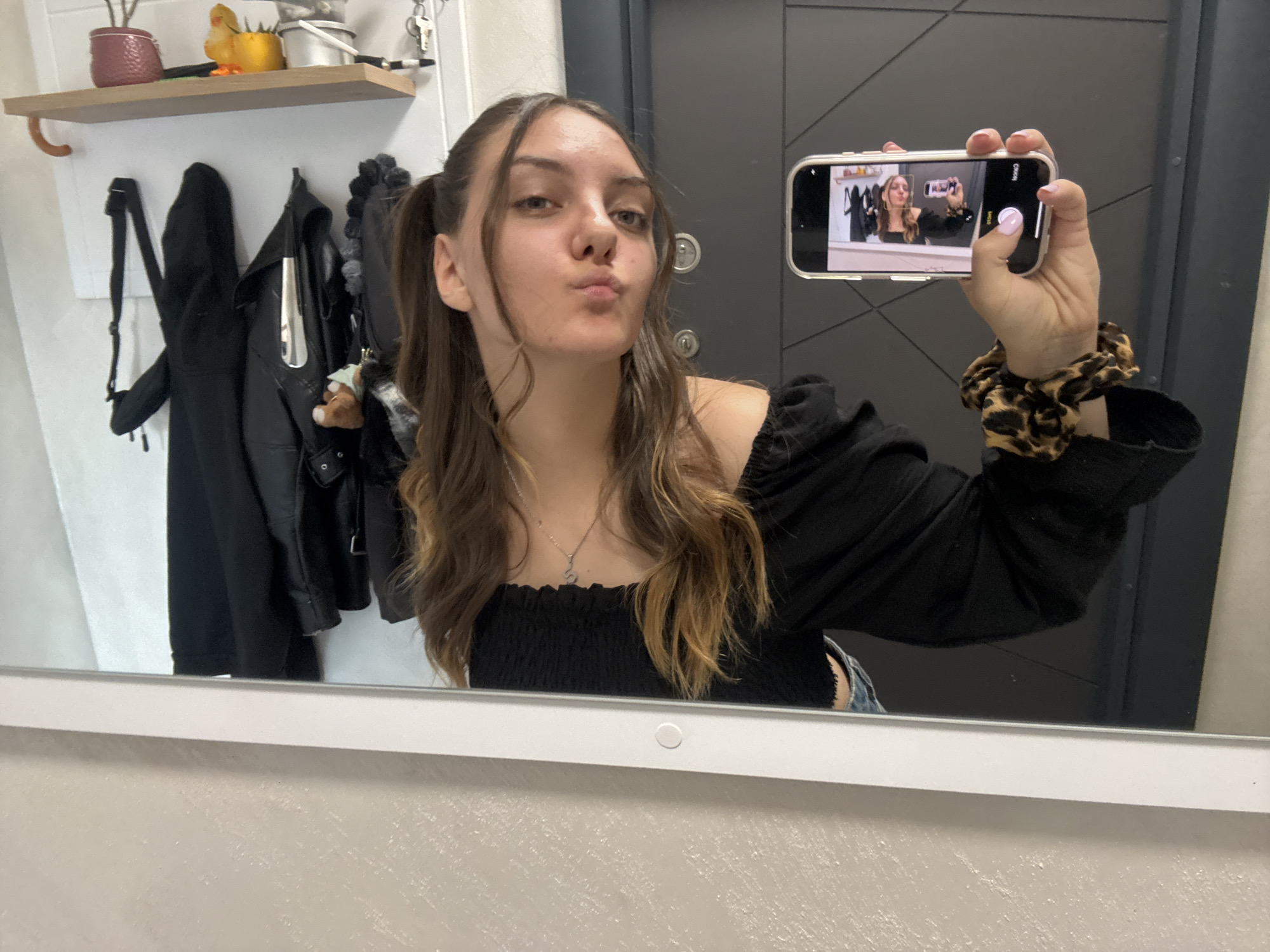 A young woman is taking a selfie in a bathroom mirror. She is holding a phone in her right hand, which reflects another image of her taking a selfie. She has brown hair in pigtails and is wearing a black shirt with a smocked bodice. The room has a shelf with various objects on it, and a coat rack with black coats. Behind her, a dark grey door can be seen. The location is likely an apartment in Sofia, Bulgaria.