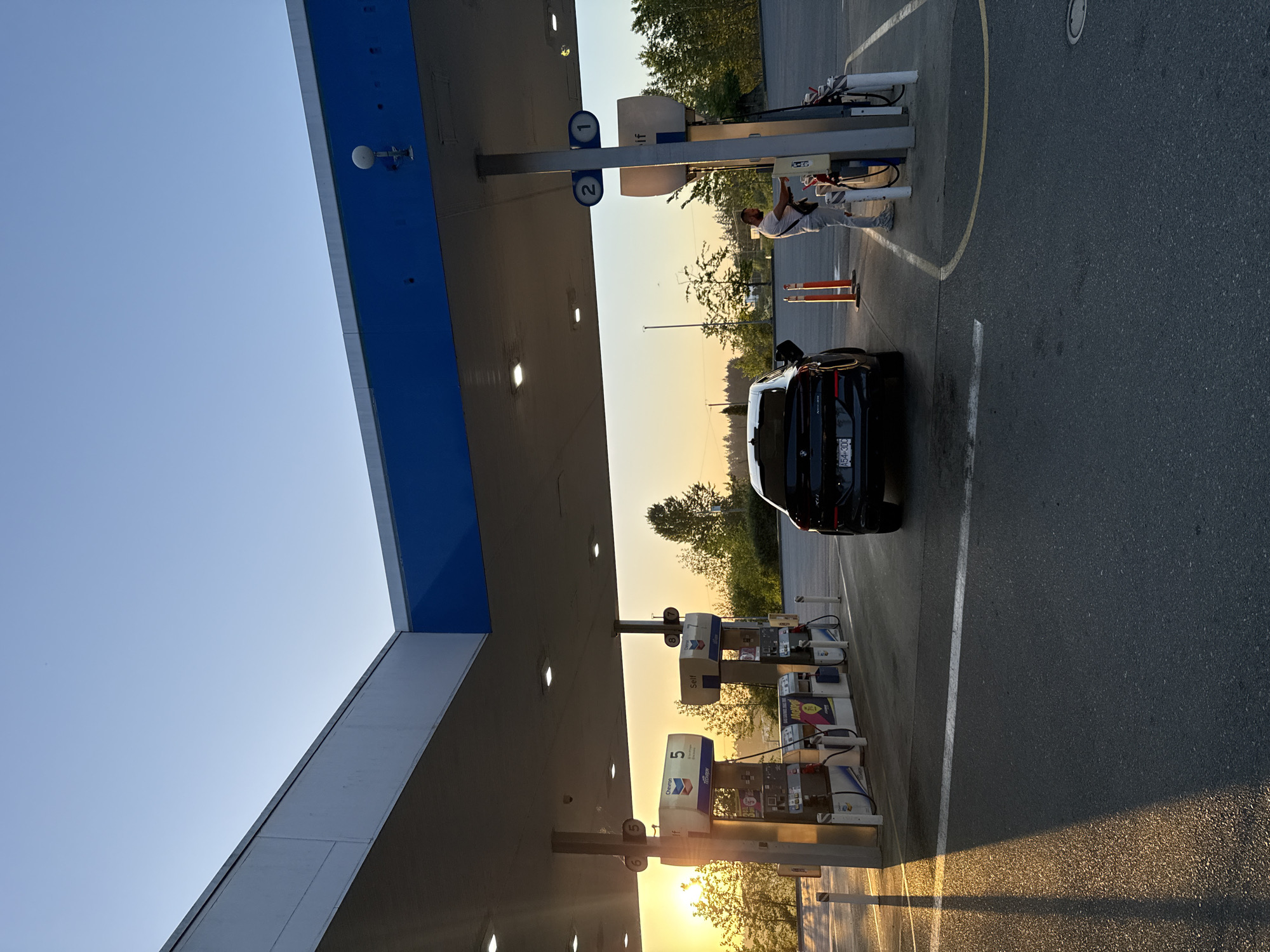 The photograph depicts a gas station in Parksville, Canada. A black car is parked at pump number 2. A person is standing near the pump on the driver's side of the car. The sun is low on the horizon, creating long shadows and a warm light. Several other gas pumps are visible in a row. The sky is clear and blue. The gas station canopy has blue accents.