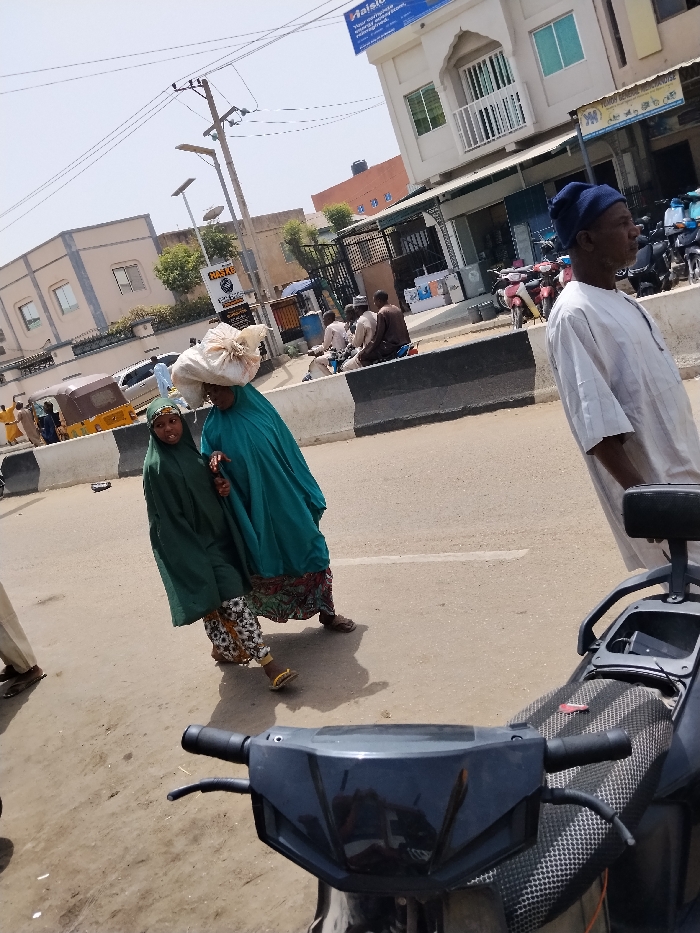 The image captures an urban street scene in Fagge D2, Nigeria, under bright daylight. In the midground, an adult female wearing a teal abaya over a patterned skirt walks from left to right, balancing a large, light-colored bundle on her head. A child, dressed in a dark green hijab and patterned skirt, walks alongside her to the left. Both wear sandals. To the right, an adult male stands, facing right, dressed in a light-colored traditional Nigerian garment and a dark blue cap. The foreground is partially obscured by the handlebars and textured black seat of a motorcycle or scooter, from which the image appears to be taken.
The paved road features a black and white concrete median barrier running horizontally across the mid-section of the frame. In the background, on the left, a light-colored auto-rickshaw (Keke NAPEP) and a white car are visible on the road, alongside another pedestrian. Further back, a multi-story building with a light-colored facade and dark window frames is present. On the right background, several men are seated on motorcycles or scooters parked along the roadside. A prominent light-colored, multi-story building with green-tinted windows, an ornate balcony, and an arched entryway is visible. A blue sign bearing the word "Haision" and text about "comfort" is affixed to its upper facade. Below it, a black and yellow sign with "HASKE" is visible. Overhead power lines crisscross the clear sky. Utility poles equipped with solar-powered streetlights are also present.