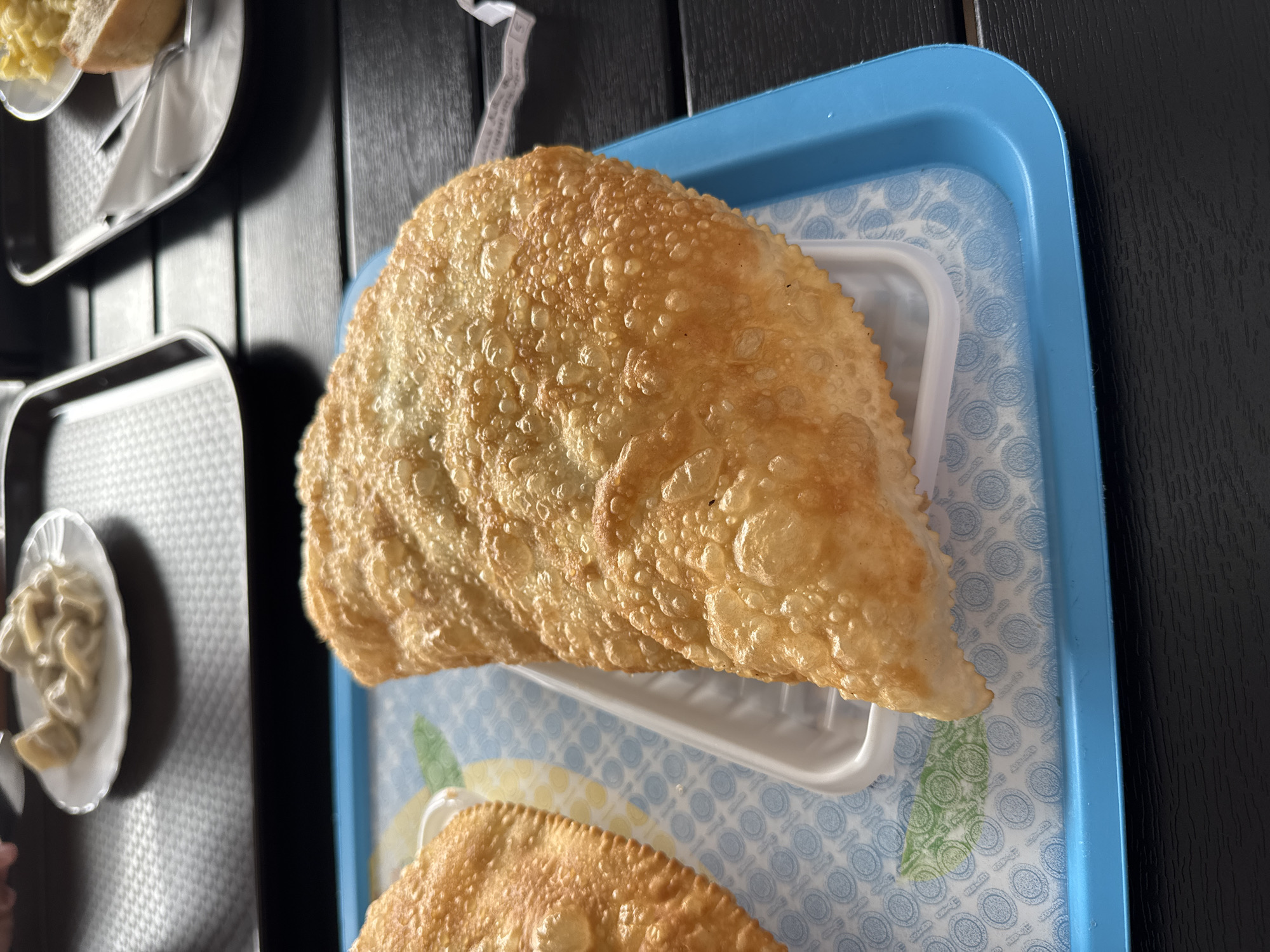 This image depicts two golden-brown chebureki, a type of deep-fried turnover, placed on a blue tray lined with patterned paper. The chebureki are semicircular and appear to be filled. The tray is situated on a dark wooden surface, likely a table, in what is presumed to be a cafe or restaurant in Novorossiysk, Russia. In the background, other food items and trays are visible, suggesting a dining setting. The lighting suggests an indoor environment with natural light.