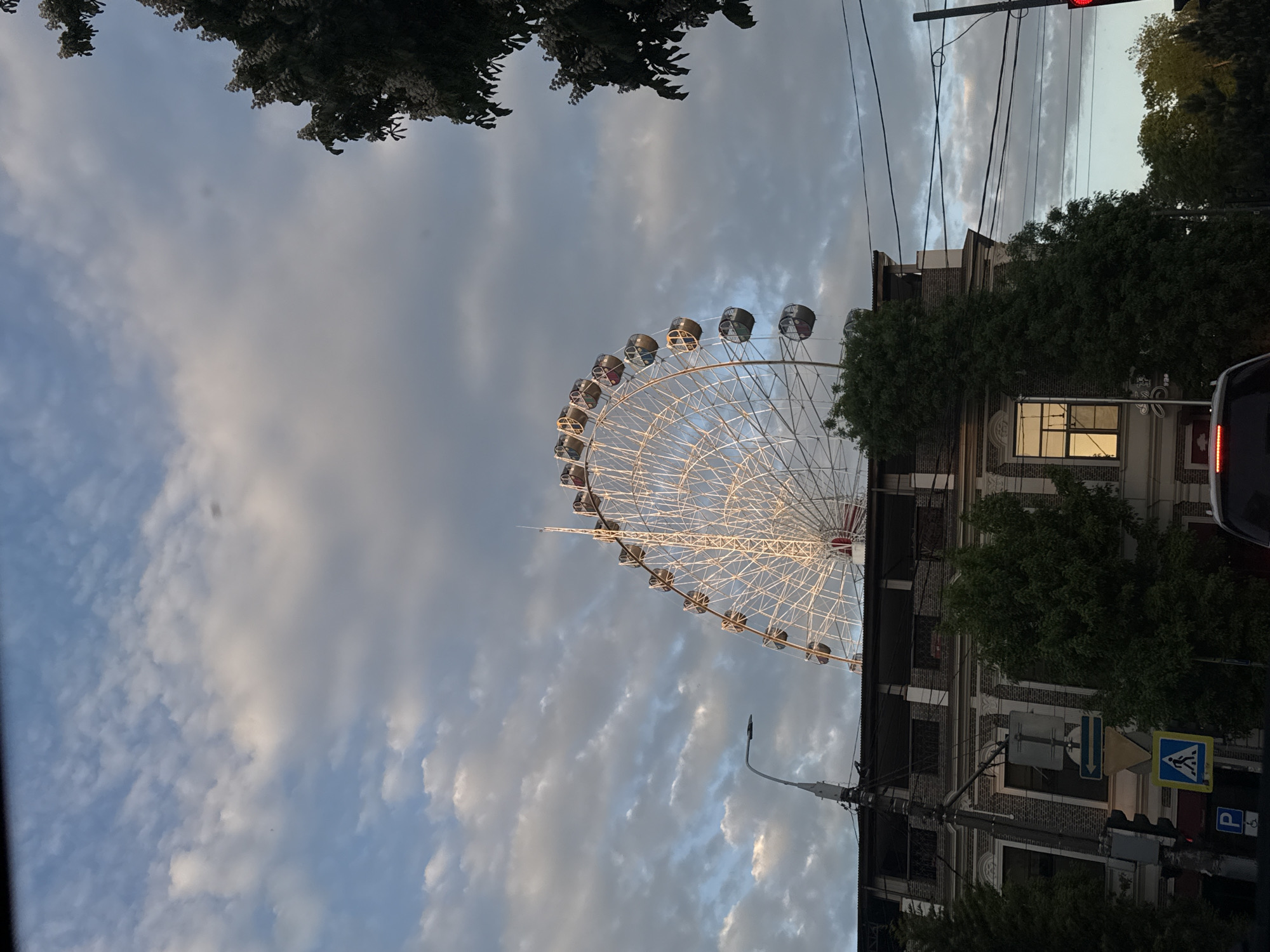 The image shows a ferris wheel positioned next to a brick building in an urban environment. The ferris wheel is white and illuminated, contrasting against a sky with a mix of blue and grey clouds. The building is partially obscured by trees, with one window emitting a warm light. A traffic light is visible at the top right corner, displaying a red light. Street signs are also visible near the building, indicating pedestrian crossing and parking. The picture is taken from a low angle, looking up towards the Ferris wheel and the building.