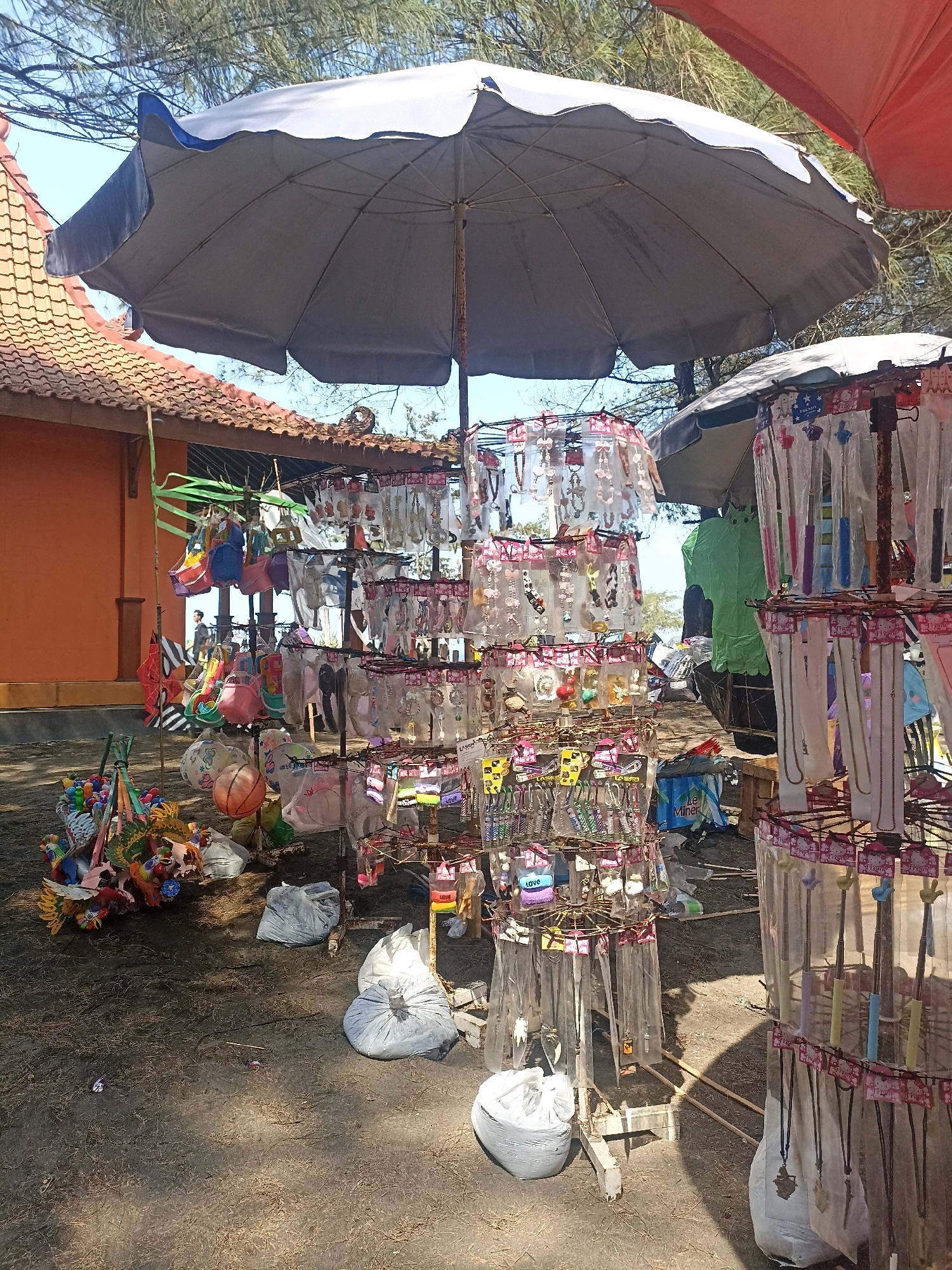In Kenoyajaya, Indonesia, a market stall stands under a large grey umbrella. The stall displays an assortment of goods, including necklaces, keychains, and small bags, all packaged and hung on rotating racks. A smaller orange building with a terracotta tiled roof is visible in the background, and pine trees with sparse foliage suggest a coastal or open area. The ground is covered in dark sand or dirt. Several large, white plastic bags are present, possibly containing unsold merchandise or refuse. The scene appears to be outdoors, under bright sunlight, casting distinct shadows on the ground.