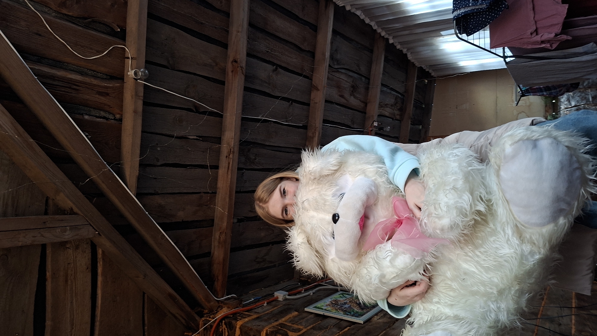 The image depicts a young woman holding a large, white, fluffy stuffed animal with a pink bow. She is wearing a light blue long-sleeved shirt and jeans. The background appears to be the interior of an attic or unfinished space with wooden walls, exposed beams, and wiring. There is also a clothes drying rack with clothing on it. The scene is presumably located in Большой Букор, Russia.
