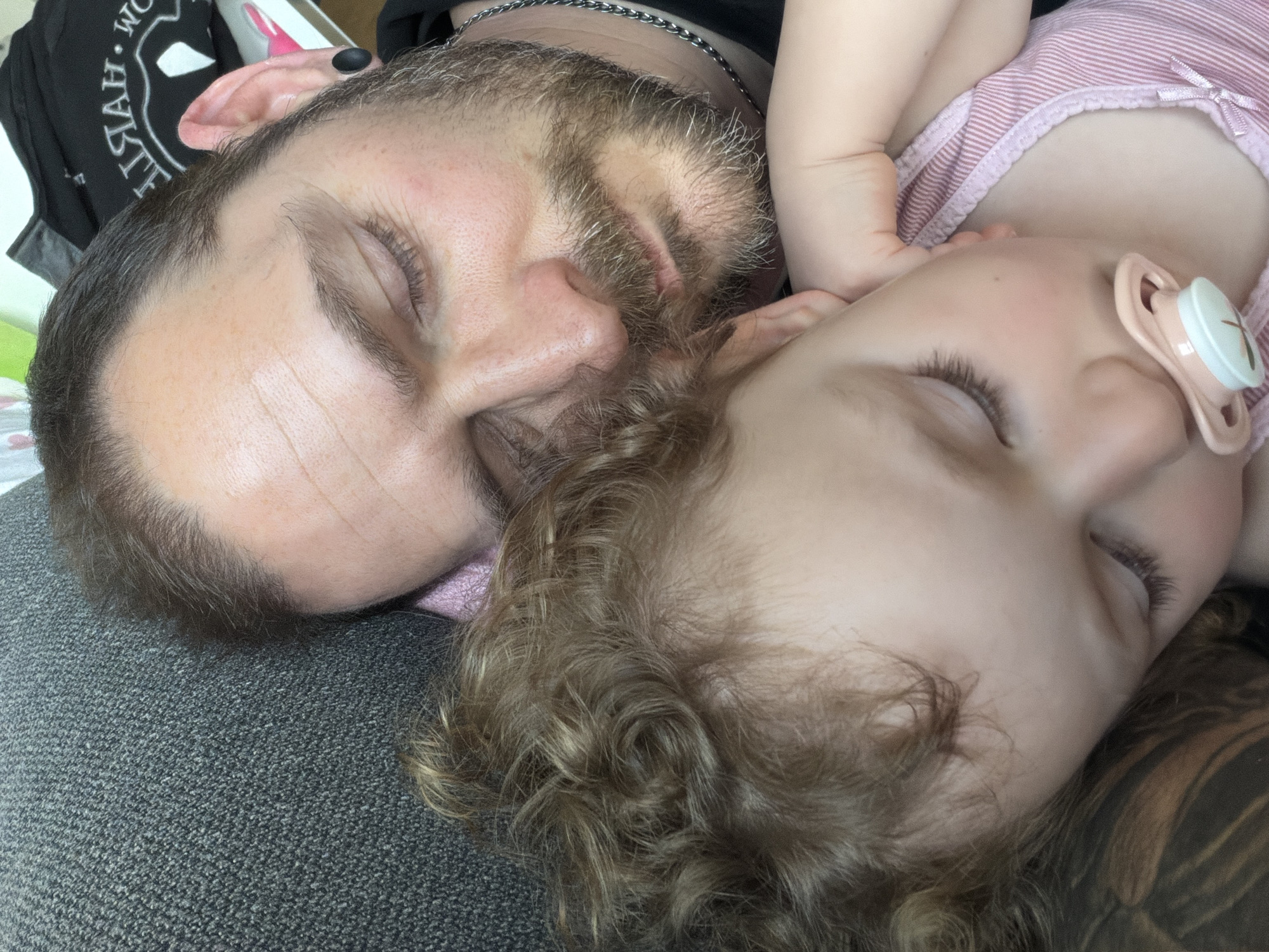 The image shows a close-up view of a man and a young child sleeping together on what appears to be a gray sofa. The man has a beard, closed eyes, and appears to have a chain necklace. The child, wearing a pink striped top, is also sleeping and has a pacifier in her mouth. The child has curly blond hair. The location and city of Trassem, Germany, cannot be discerned from this image.