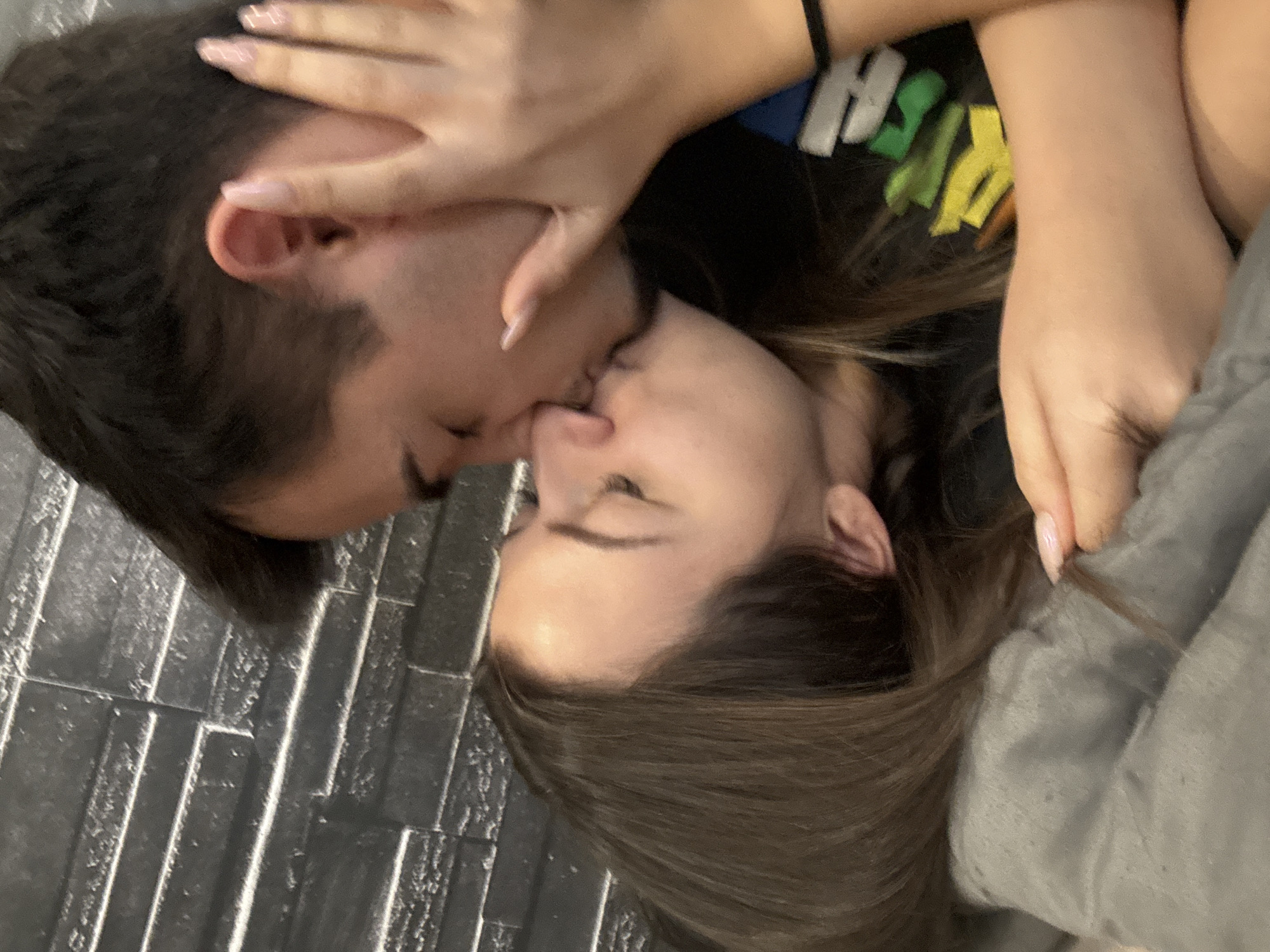 A man and woman are lying on a tiled surface, face to face, and kissing. The man is on the left, with dark hair. The woman is on the right, with brown hair. A hand is touching the man's head. The woman is wearing a black top with colorful lettering. The setting appears to be indoors. Location details are unavailable.