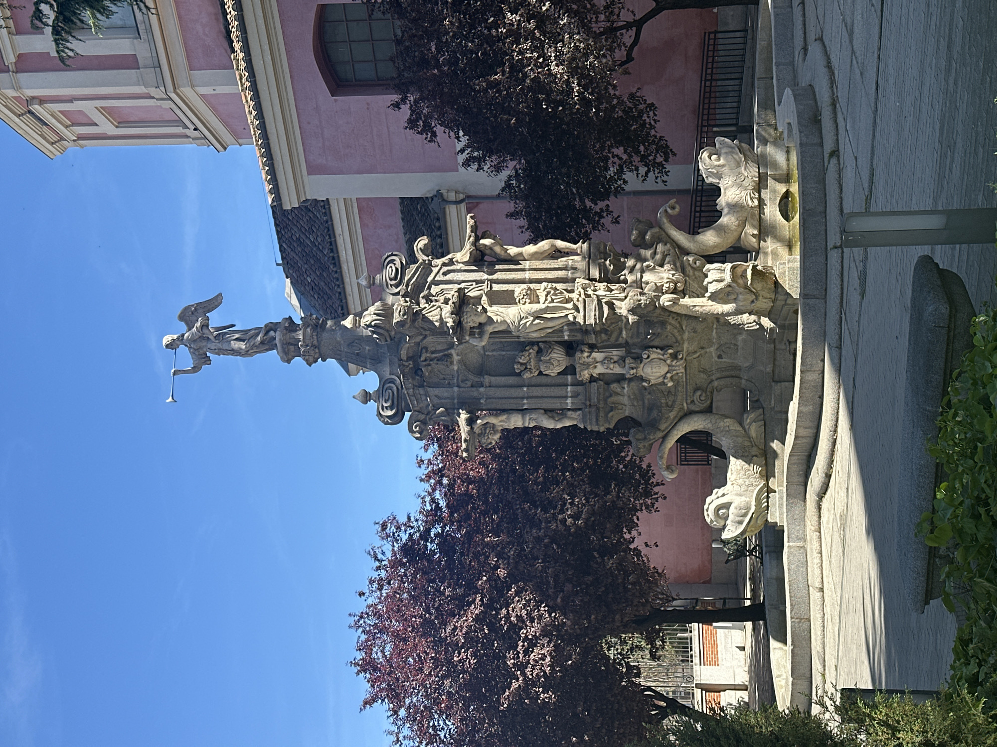 The image depicts the Fountain of Fame (Fuente de la Fama) in Madrid, Spain. The fountain is made of light-colored stone and features an elaborate sculptural design. It includes figures of mythological creatures and a statue of Fame atop a tall column, holding a trumpet. The fountain is surrounded by trees with reddish leaves and a pink building with a window in the background. The sky is blue. The setting appears to be a public square or plaza. The statue of Fame is central to the composition.