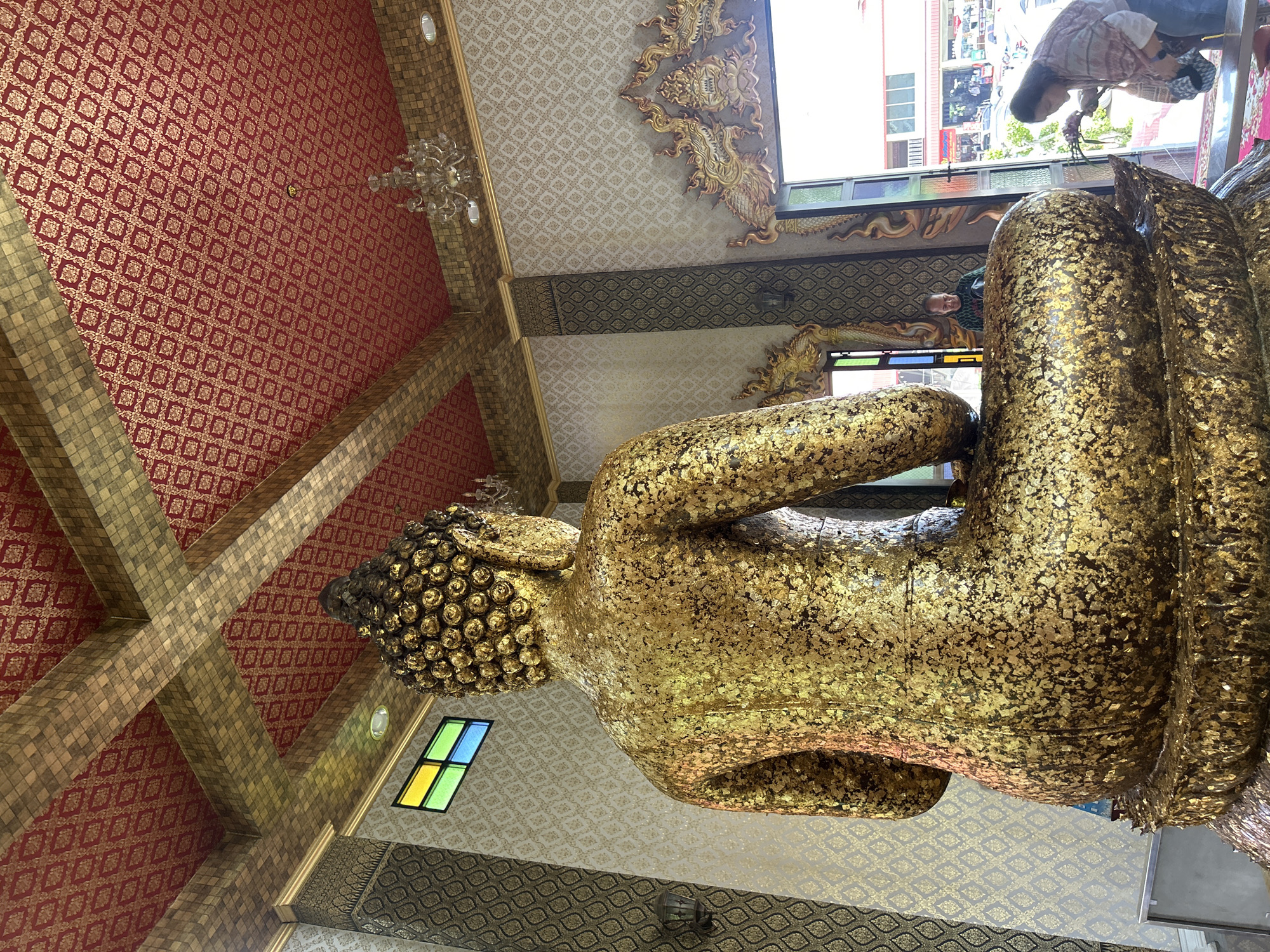 In Thai Ko Subdistrict, Thailand, an interior view of a temple features a large, golden Buddha statue, predominantly covered in gold leaf, seated in a meditative pose. The statue's back is facing the camera, revealing the intricate details of its robes and the textured surface of the gold leaf. The surrounding environment is elaborately decorated with ornate, gold-colored architectural elements and patterned wallpaper. The ceiling is adorned with red fabric featuring intricate gold patterns and suspended golden beams, along with a crystal chandelier. A stained-glass window with green, yellow, and blue panes is visible on the left. In the background, through an open doorway, a street scene with buildings and parked vehicles can be observed. Two individuals are present: one man stands near a mirrored surface on the left, and a woman is seen in the foreground on the right, appearing to offer something to the Buddha statue.