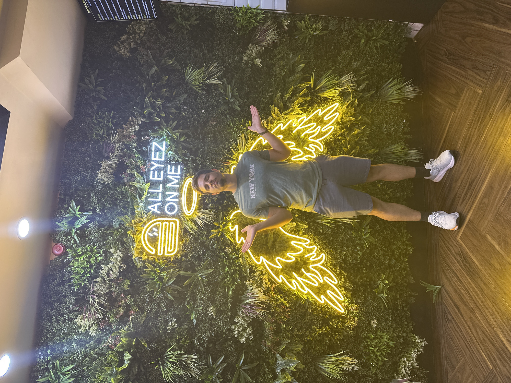 A man stands in front of a living wall decorated with a yellow neon sign that reads "ALL EYEZ ON ME" above a hamburger icon and angel wings. The man is wearing a green t-shirt with "NEW YORK" printed on it, grey shorts, white sneakers, and black socks. He has his arms outstretched and is looking towards the camera with his eyes closed. The living wall is made of various green plants and is located indoors, as indicated by the ceiling lights. The floor is made of dark wood laid in a chevron pattern. A menu board is partially visible on the right side of the image. The location is identified as a burger restaurant in Leeds, United Kingdom.