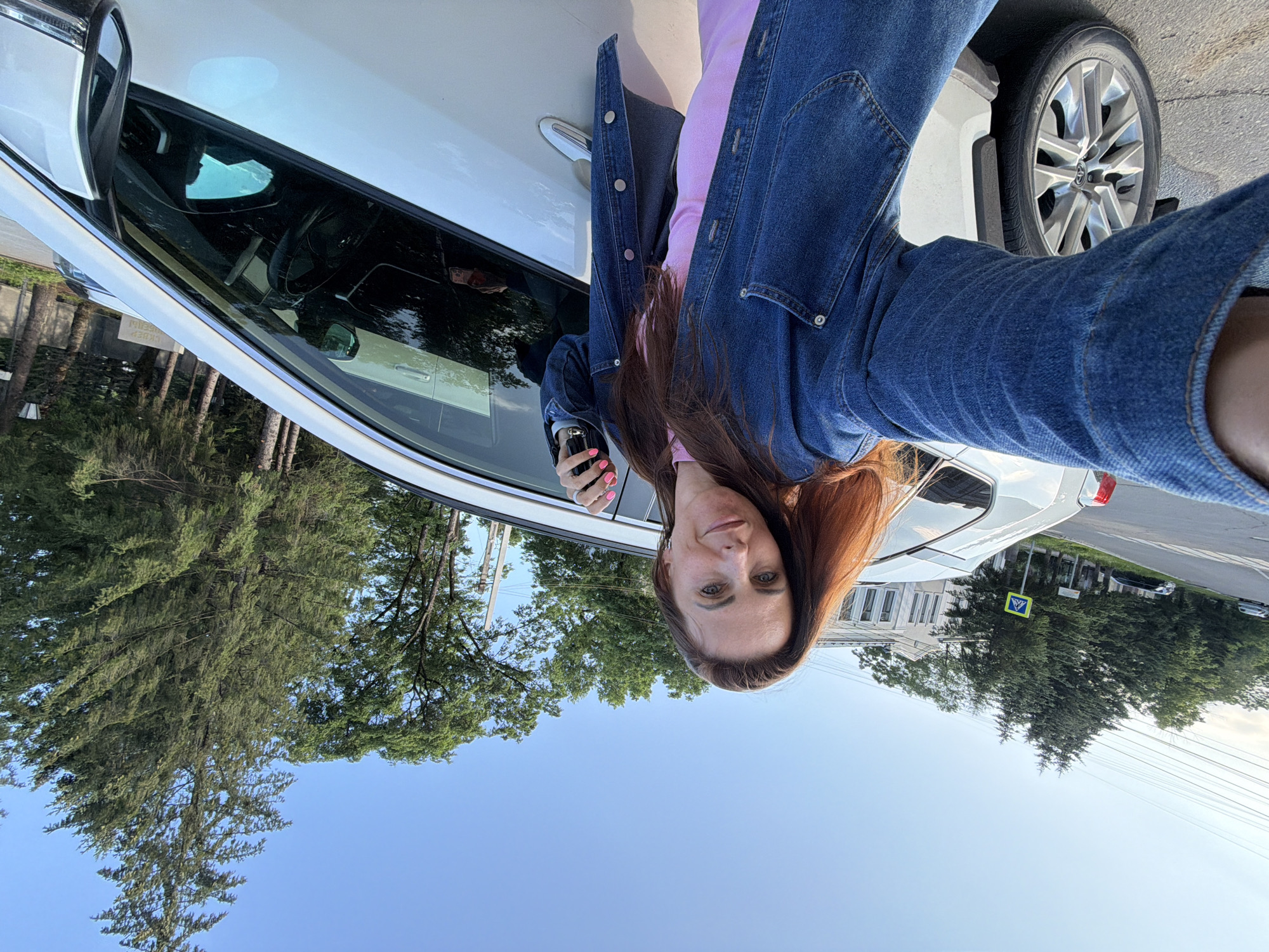 The image shows a woman taking a selfie next to a white car. She has long, reddish-brown hair and is wearing a pink top and a denim jacket. She is holding a car key in her right hand. The car door is open, and the interior is visible. The background shows trees and a building, suggesting an outdoor setting, possibly a street or parking area. The sky is blue with some wispy clouds, indicating it is daytime, likely late afternoon or early evening due to the warm lighting. There is a pedestrian crossing sign visible in the distance. The image appears to be taken in Birobidzhan, Russia. The overall mood seems casual and relaxed.