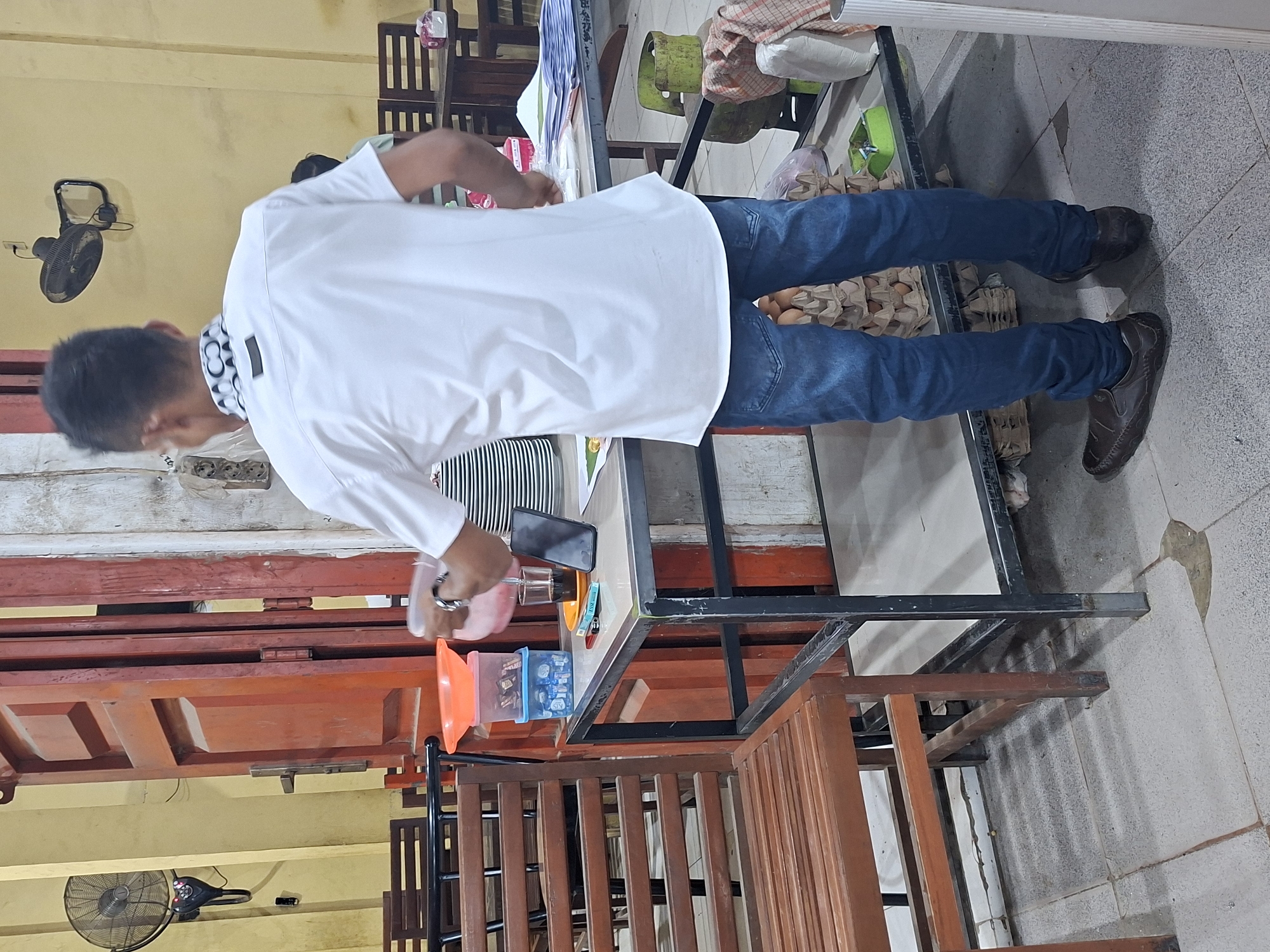 The image shows a person in a white shirt and blue jeans standing at a table in what appears to be a food establishment in Keudegeudong, Indonesia. The person is holding a plastic container and pouring something into a glass. On the table, there is a smartphone, a lighter, a stack of plates, and some papers. In the background, there are wooden chairs and tables, a red door, a wall-mounted fan, and some stacked items including egg cartons. The lighting suggests an indoor setting, possibly a warung or small restaurant.