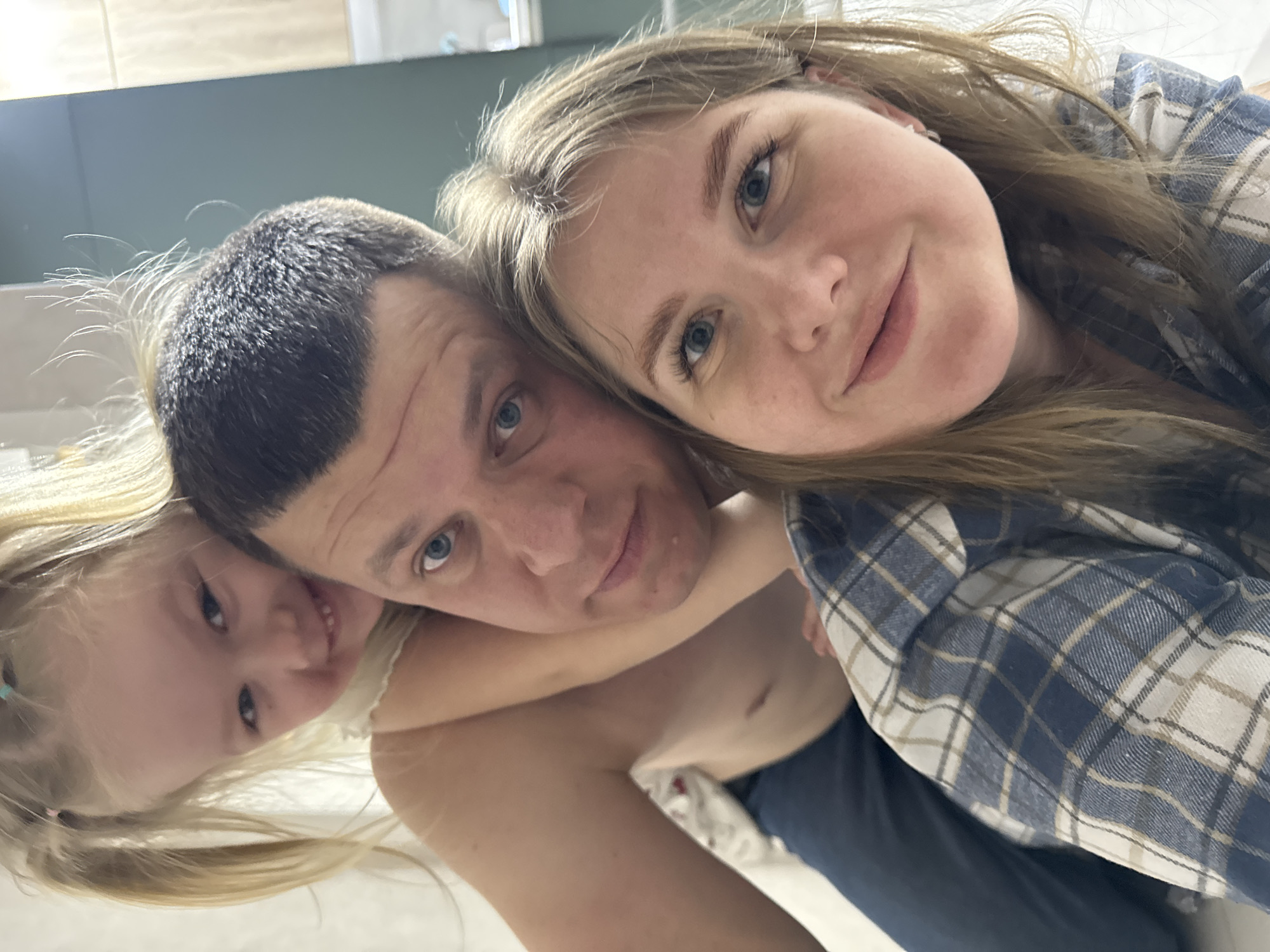The image depicts a family selfie taken indoors. A fair-skinned man with short, dark hair is in the center, looking directly at the camera. He has blue eyes and a neutral expression. To his right, a fair-skinned woman with long, blonde hair is leaning her head against his. She is wearing a blue and white plaid shirt and is also looking at the camera with a slight smile. A young, fair-skinned girl with long, blonde hair is to the left of the man, smiling into the camera. She has her arm wrapped around the man's neck. The background includes light-colored tile. The location is reported to be in Gornyi (Dvurechenskoe S/P), Russia, but this cannot be verified from the image itself.