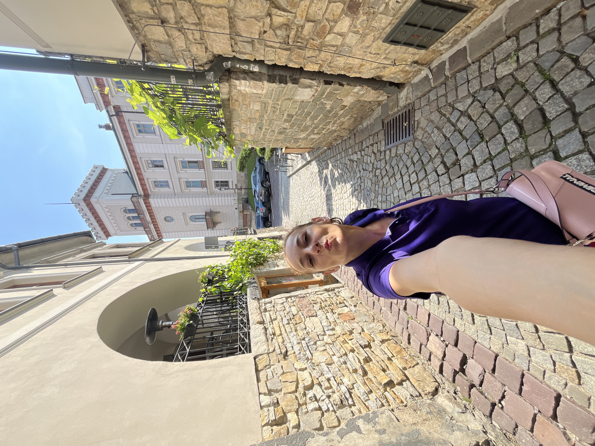 In an outdoor scene, a fair-skinned woman with auburn hair pulled back is taking a selfie while walking down a narrow cobblestone alley. She is wearing a vibrant purple polo shirt and a light pink handbag is slung over her right shoulder. The alley walls are constructed of rough-hewn stone blocks. On the left, a building features a pale beige facade with an arched walkway. On the right, a building with a similar stone facade has a downspout running vertically along its surface. In the background, a large, light-colored building with a distinctive crenellated tower rises, accompanied by a clear blue sky. Several parked cars are visible further down the alley. The lighting suggests it is daytime.