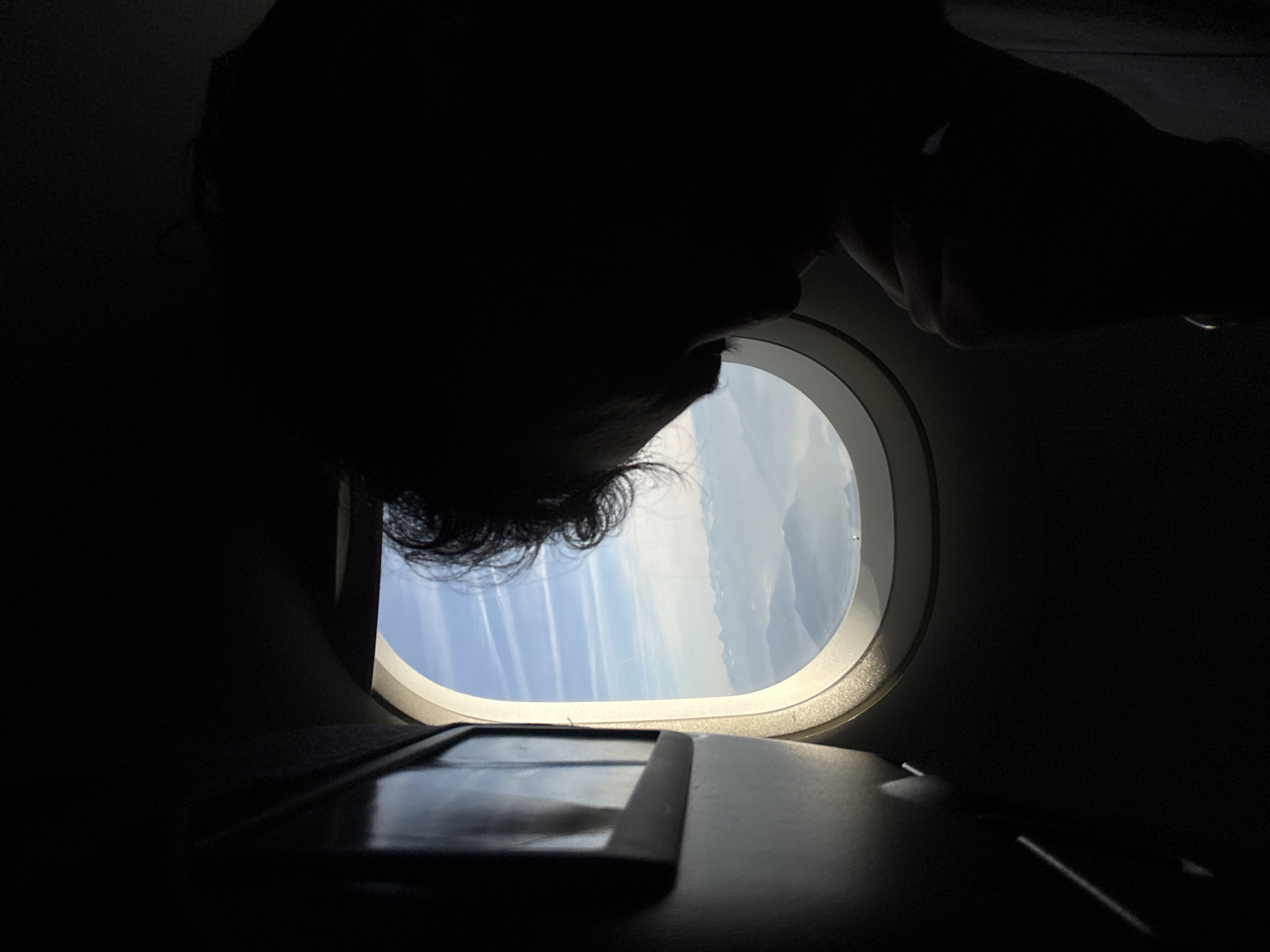 The photograph shows a dark, low-angle interior shot from inside an airplane, likely at cruising altitude. A person's head and hand are silhouetted against the bright light coming through the plane window. Outside the window, a blue sky with streaking clouds can be seen. Below the window, there's a partially visible tray table with an electronic device on it. The location is not discernible from the content of the photograph. It is not possible to determine if the location is Tradate, Italy, based on this photograph.