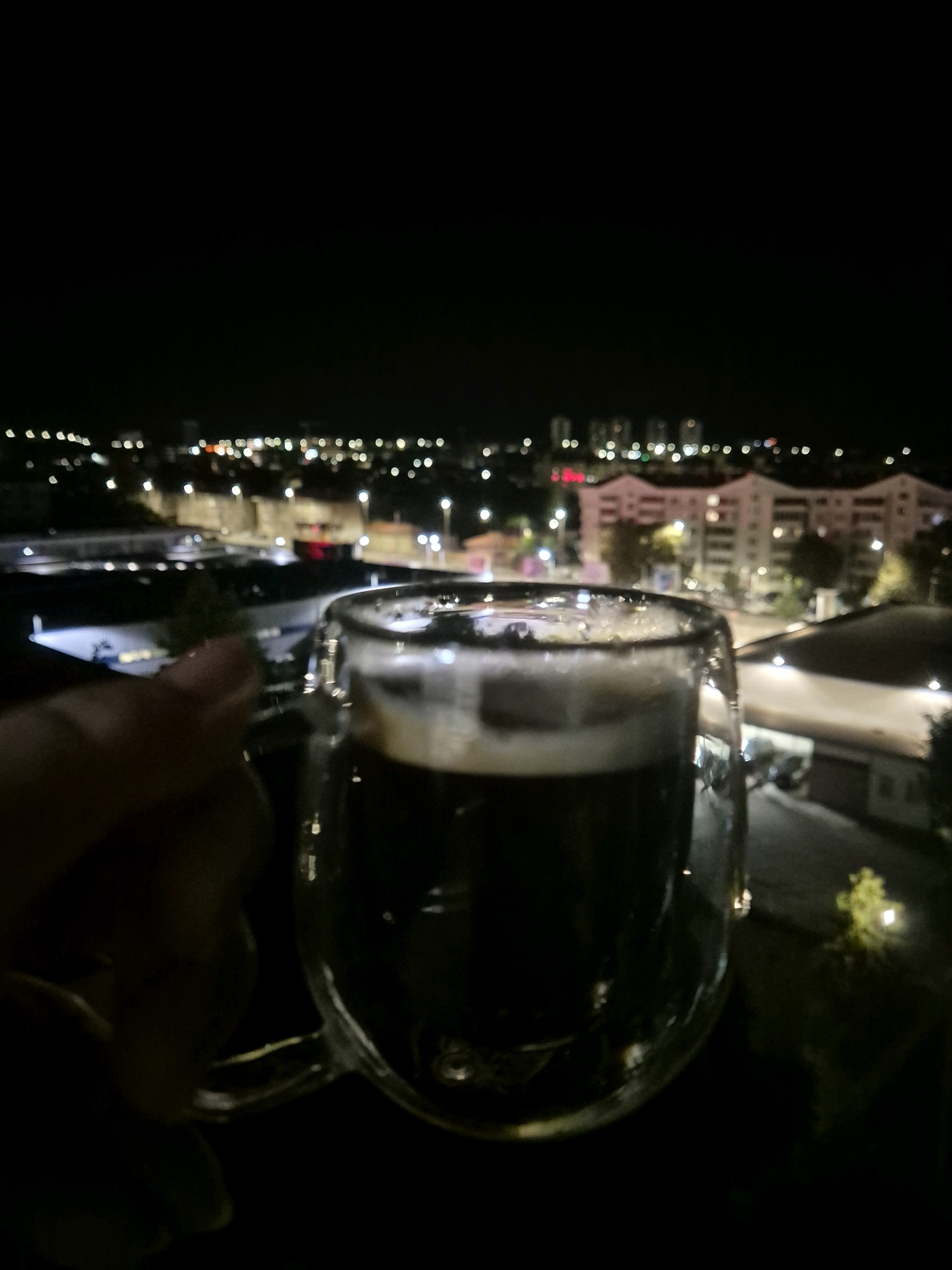 In the foreground, a hand is holding a clear glass mug containing a dark liquid with a foamy top, likely a beverage like coffee or beer. The mug has a double-walled design. In the background, a blurry cityscape is visible at night, with illuminated buildings and streetlights creating bokeh effects. The scene appears to be an outdoor setting, possibly a balcony or rooftop, overlooking a residential or urban area.