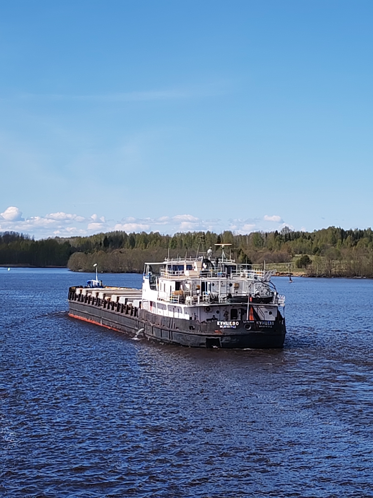 The image depicts a black and white barge labeled "КУНЦЕВО" (KUNTSEVO) on a wide body of water, likely a river. The barge is oriented away from the viewer, moving through the water and creating a small wake. The river is flanked by a tree-lined bank and a clear blue sky with scattered clouds.
The barge is in Pruzhinino, Russia.