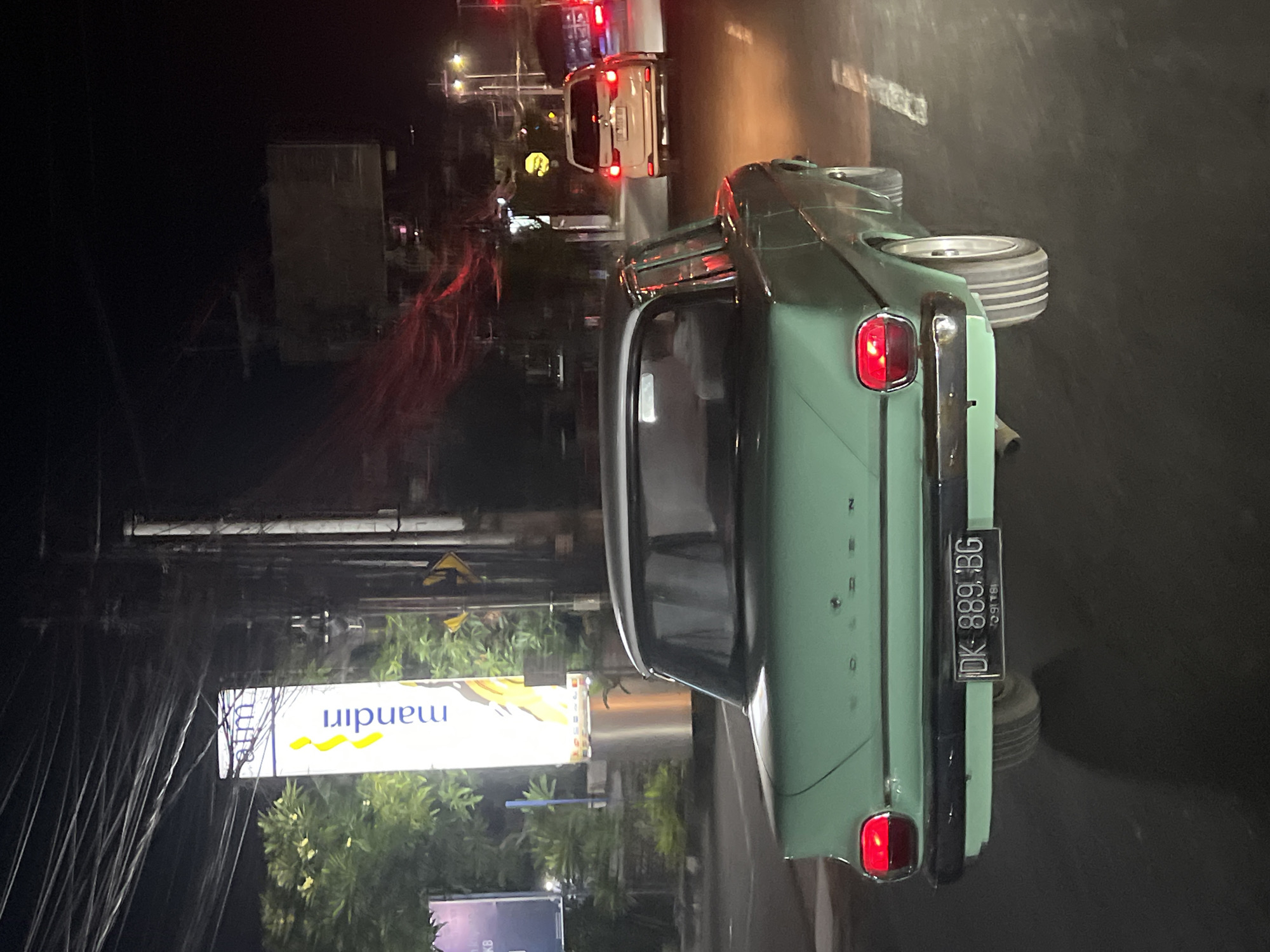 A long-shot photo shows a light green vehicle parked on a street in Denpasar, Indonesia. The license plate number is DK 889 BG. A Mandiri sign is visible. There are utility poles and wires running alongside the street. Other cars are on the street, and it is dark.