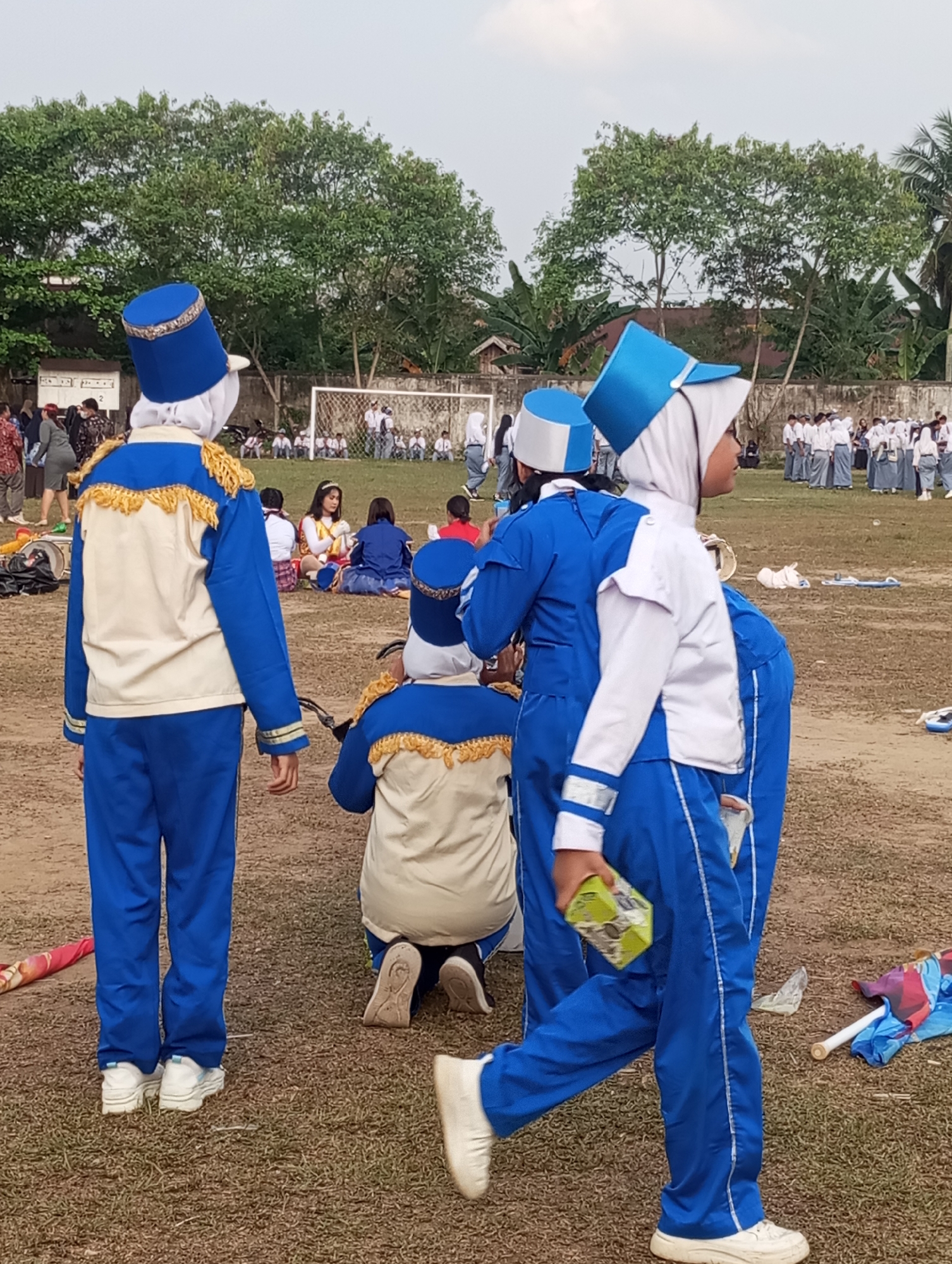 In Candirejo, Indonesia, a group of individuals in blue and cream marching band uniforms are gathered on a grassy field. Two individuals in the foreground are facing away from the camera, wearing blue hats with white bands and ornate gold trim on their jackets. One of them is holding a small green box. Further in the background, other individuals in similar uniforms are visible, some sitting on the ground. A soccer goal is present on the field, and trees line the perimeter of the area. The sky is overcast.