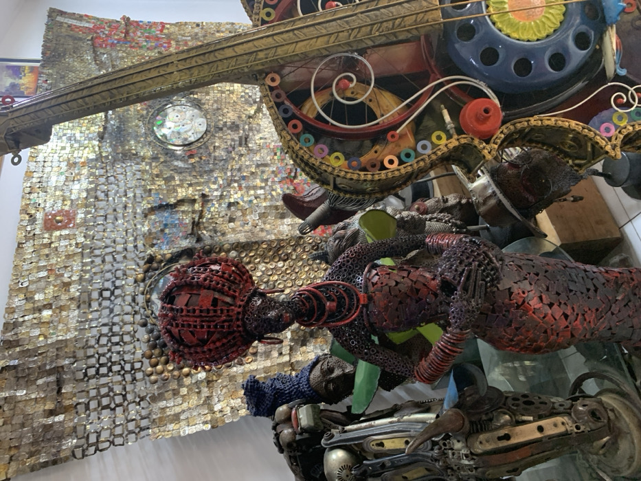 The scene, an interior art display located in Ikate, Nigeria, features an array of sculptures crafted from repurposed materials. A prominent, tall female sculpture, constructed from interconnected dark red metal components, stands centrally. This figure displays an elaborate crown, multi-strand necklaces, and bangles, all consistent in material and dark red hue. To her immediate left, a partially visible, abstract humanoid sculpture is present, fashioned from various metallic parts and topped with a blue knitted headpiece. The background is dominated by a large wall mosaic composed of numerous flattened metallic rectangles, likely recycled can materials, creating a textured, shimmering surface. This wall art includes irregular dark patches and a circular element containing embedded compact discs. To the right, a large, stylized string instrument, resembling a cello or double bass, is visible. This instrument is assembled from metal, bicycle chains, gears, and colorful rings, with a blue telephone dial integrated into its body. A smaller, dark-toned head sculpture wearing a fedora-style hat is partially visible at the base of the string instrument. Sections of white wall are visible on the left and in the upper background.