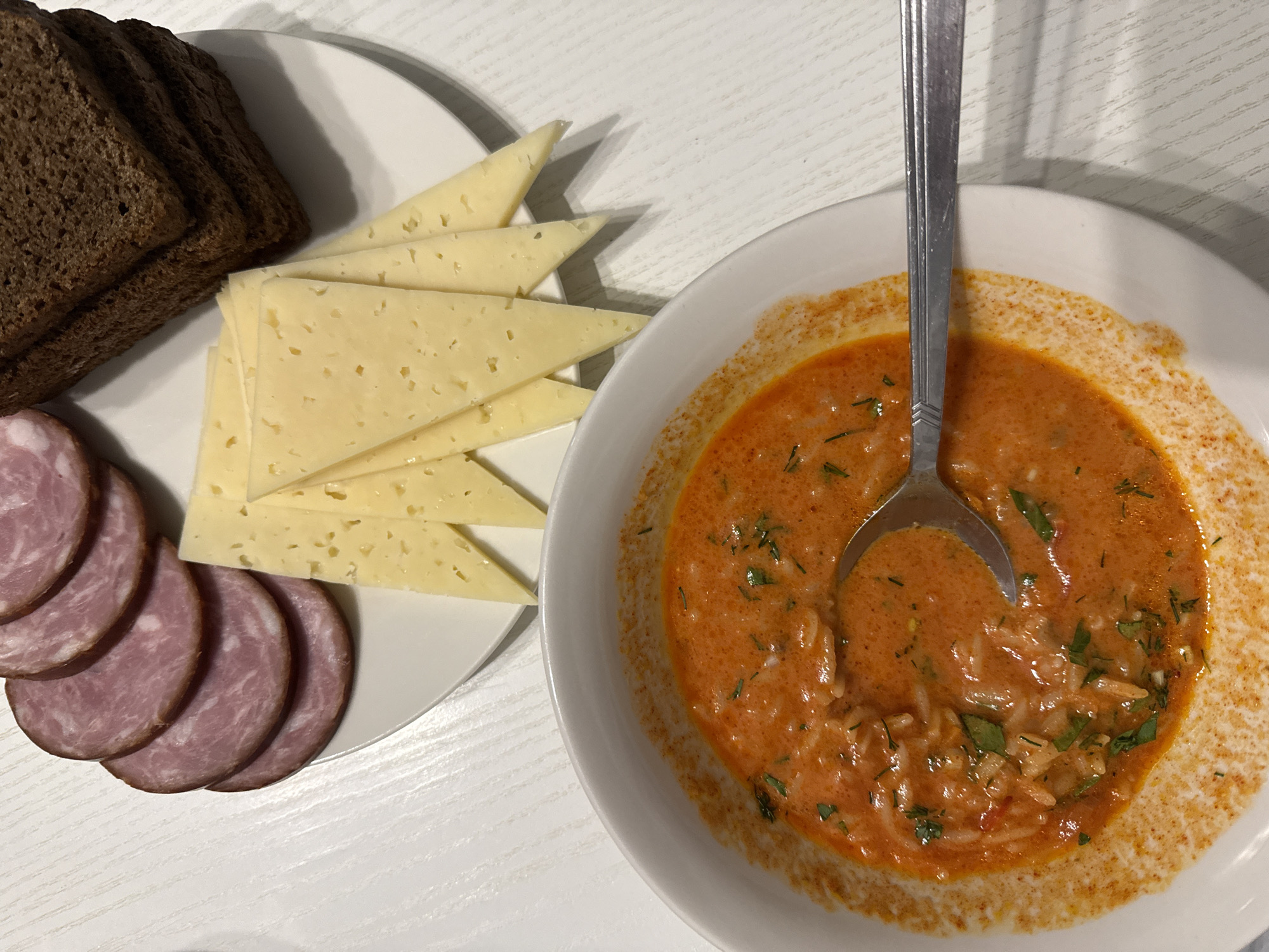 The image depicts a meal consisting of a bowl of soup, slices of cheese and sausage, and bread on a white table. A spoon rests inside the bowl of soup, which appears to have a creamy, orange base with herbs. The cheese is yellow with holes, and the sausage is pink. The bread is dark and appears to be rye. The location is unknown.