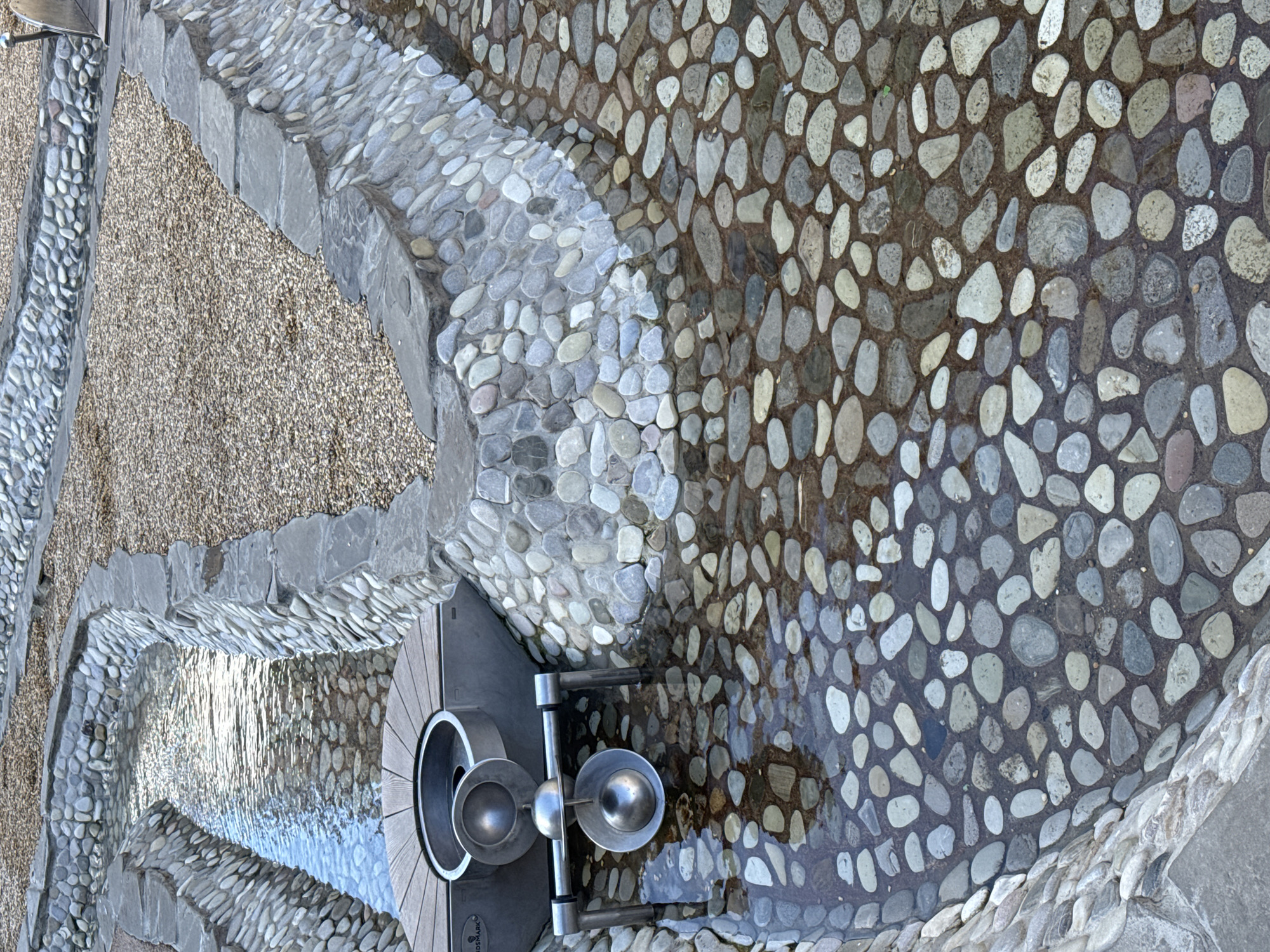 The image depicts a water feature in Novoselye, Russia, designed with various stone elements. A shallow stream of water flows through a channel lined with smooth, rounded river stones. The channel itself is bordered by a more structured stone wall constructed from larger, flat grey stones on top and a mosaic of river stones below. To the left of the water channel, a wooden platform encircles a circular metal basin. Below this platform, a metallic water play structure is visible, featuring two hemispherical cups connected by a horizontal bar and a central spherical weight suspended in water. The ground surrounding the stone walls is covered with small, light brown gravel. The overall scene suggests an outdoor recreational area, possibly a park or playground, with a focus on interactive water elements.