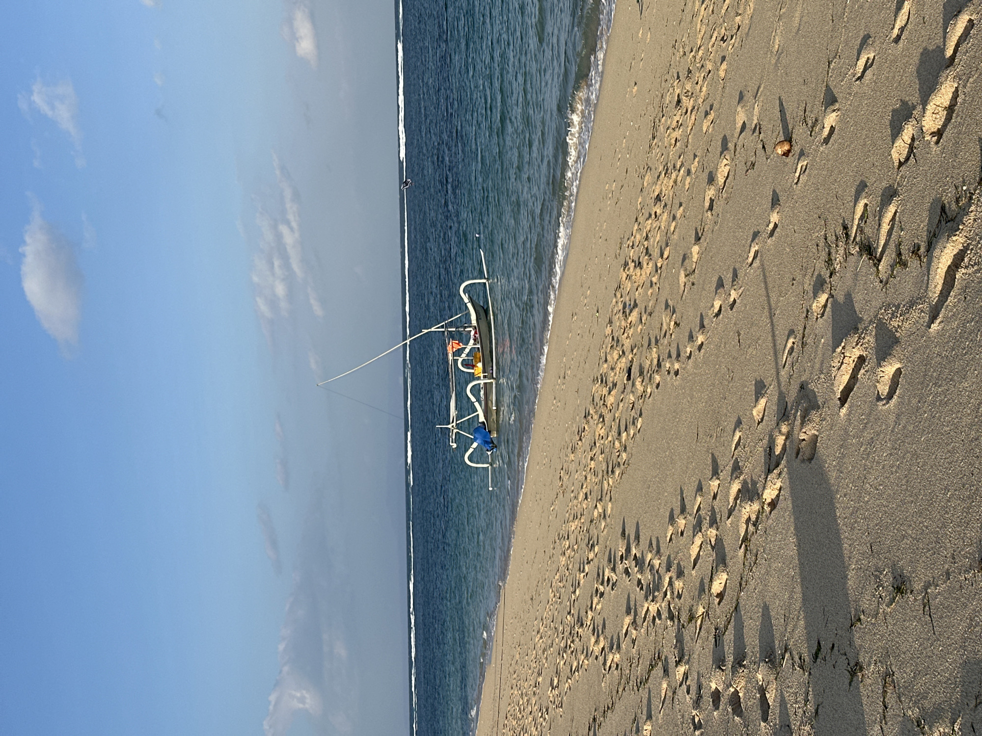 The image depicts a tranquil coastal scene in Kuta Selatan, Indonesia, likely during early morning or late afternoon, characterized by soft, warm light casting elongated shadows on the sand. The upper portion of the frame features a clear blue sky with scattered cumulus clouds. Below the sky, the vast expanse of the calm, blue-green ocean extends, with multiple lines of white-capped waves visible in the distance, indicating an offshore reef or sandbar.
In the mid-ground, a single traditional Indonesian outrigger boat, likely a jukung, is partially submerged in the shallow water near the shoreline. The boat is predominantly white with a prominent mast-like structure and distinct curved outrigger pontoons. A blue tarp or similar item is secured on the boat's interior, and a small orange object is also visible. The boat appears to be either anchored or temporarily beached.
The foreground is dominated by a wide, light-colored sandy beach, stretching diagonally from the lower left to the upper right. The sand is densely covered with numerous human footprints, varying in depth and direction, indicating recent pedestrian traffic. Interspersed among the footprints are small patches of dark green seaweed or organic debris. In the lower right quadrant of the sand, a small, round, brown object, possibly a coconut or fruit, rests near the water's edge. No individuals are visible within the frame.
