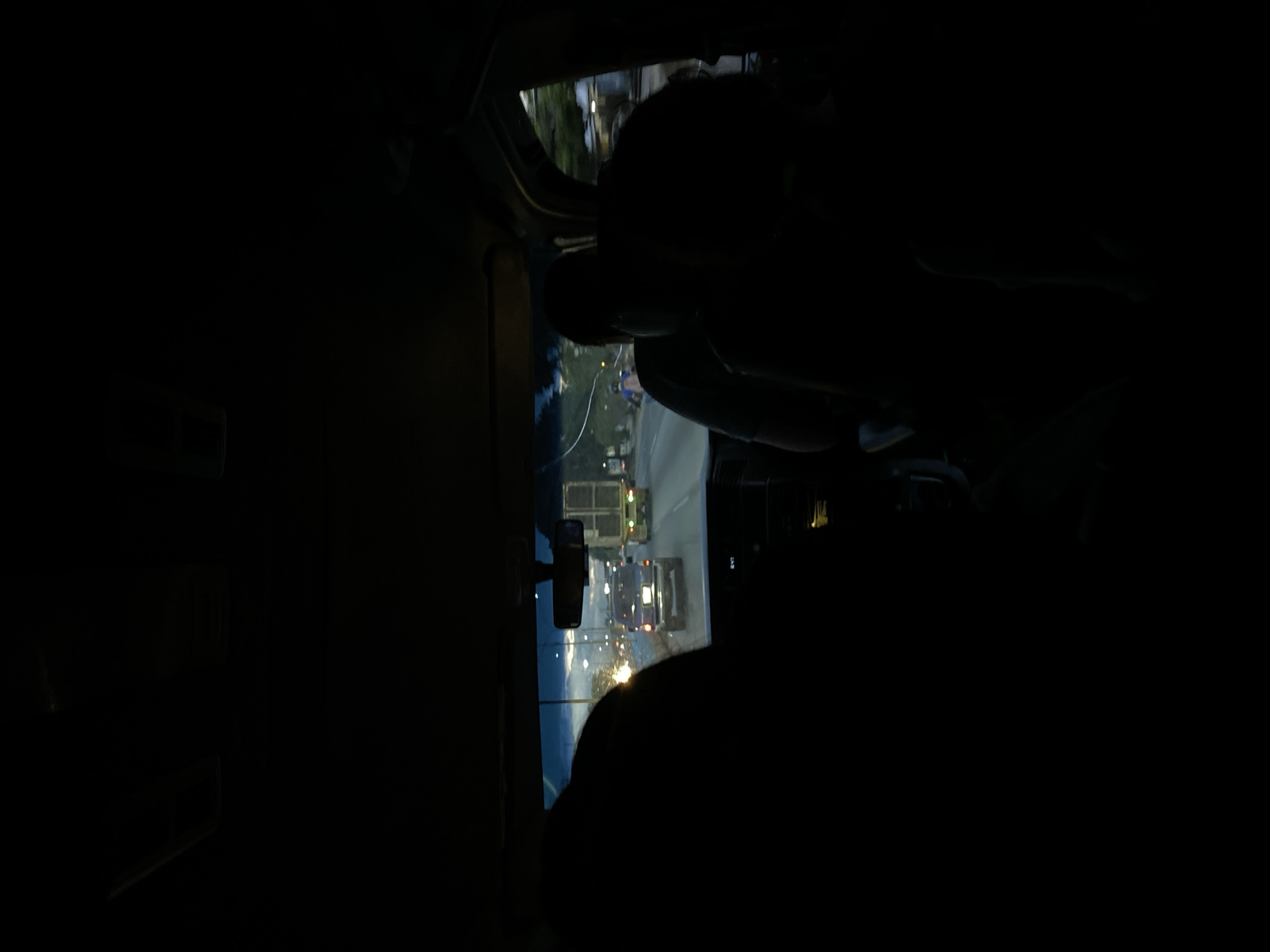 The image displays a night-time scene viewed from inside a vehicle, looking out through the windshield. The foreground is dominated by the dark silhouettes of at least two occupants, likely a driver and a passenger, with one prominent head/upper body silhouette visible on the right side. A rearview mirror is positioned centrally at the top of the windshield.
Outside the vehicle, the scene is dark, illuminated by various artificial lights. Several other vehicles, including what appear to be trucks, are visible ahead on a road or highway, their lights creating bright points. Distant streetlights or building lights contribute to the illumination, casting reflections or glare on the windshield. The setting appears to be an urban or peri-urban area, consistent with traveling through Tocuyito, Venezuela, at night. The activity depicted is driving or being a passenger in a vehicle during nighttime. No specific weather conditions are discernible, nor is any legible text visible within the frame.