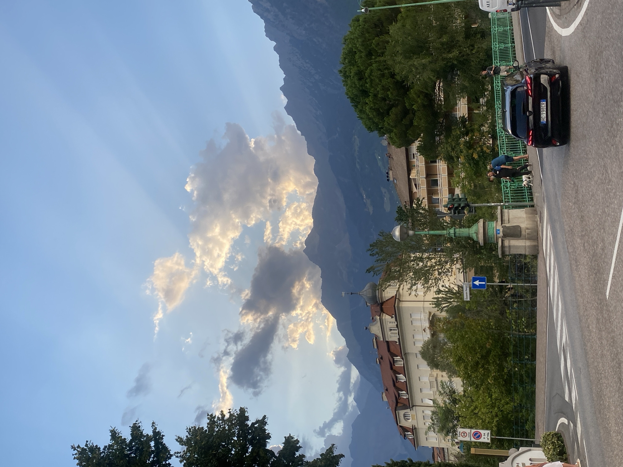 The image shows a street scene in Meran - Merano, Italy, with a view of mountains in the background. A black car with license plate HC 198CR is parked on the right side of the road, facing away from the camera. Two people are standing near the car, and a small white dog is on a leash. A green bridge with a railing spans the road in the middle of the image. On the left side of the road, there is a building with a red roof and a tower. The sky is partly cloudy, with rays of sunlight breaking through the clouds. There are trees and streetlights visible in the scene.