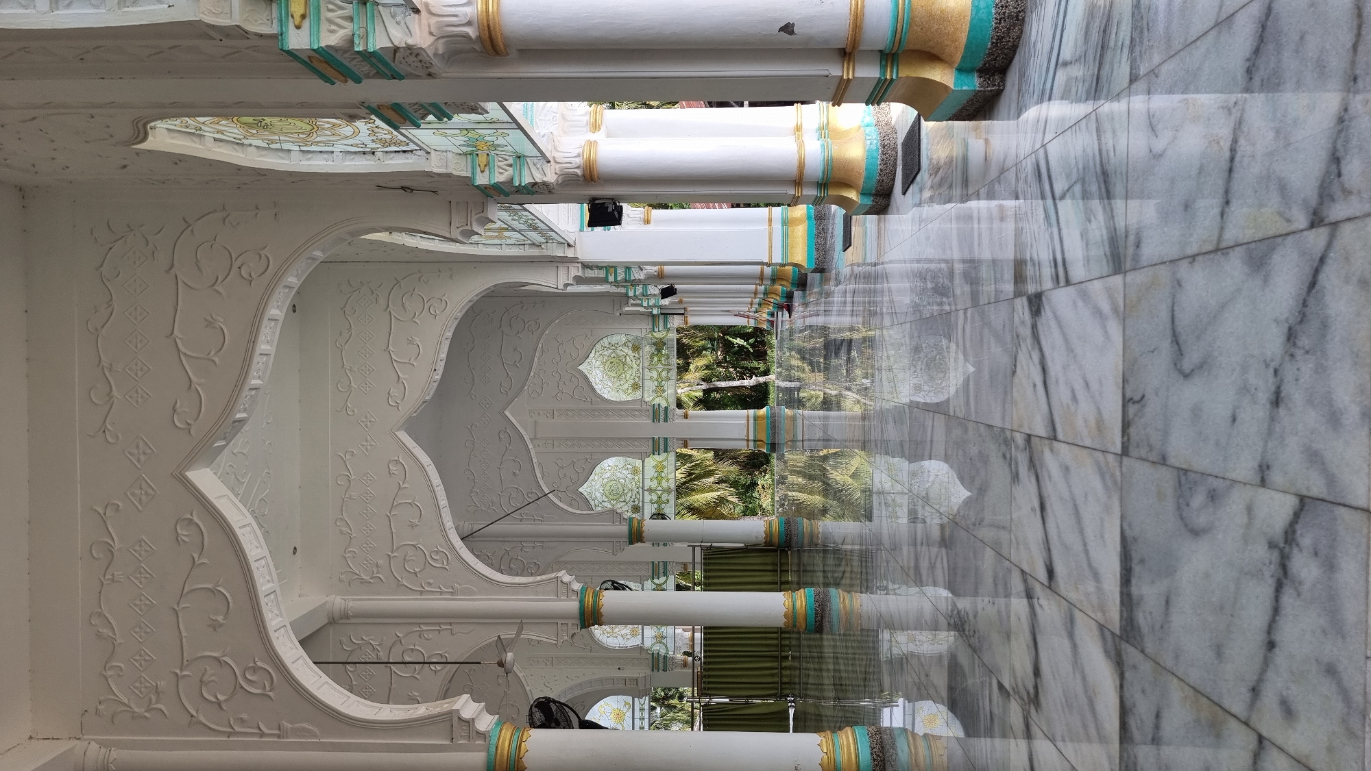 The image depicts the interior of a mosque with a focus on its architectural features and reflective marble floor. The scene is set in the city of Lapang Timu, Indonesia. The mosque features a series of white columns with ornate gold and teal detailing at their bases and tops. These columns support arched walkways with intricate white relief carvings of floral and geometric patterns. The arches lead to a view of lush green tropical foliage outside, visible through decorative, stained-glass-like window panels within some of the arches. The marble floor is highly polished and reflects the columns, arches, and the outdoor scenery, creating a sense of depth and grandeur. No people are visible in the image. The overall impression is one of serenity and architectural beauty.